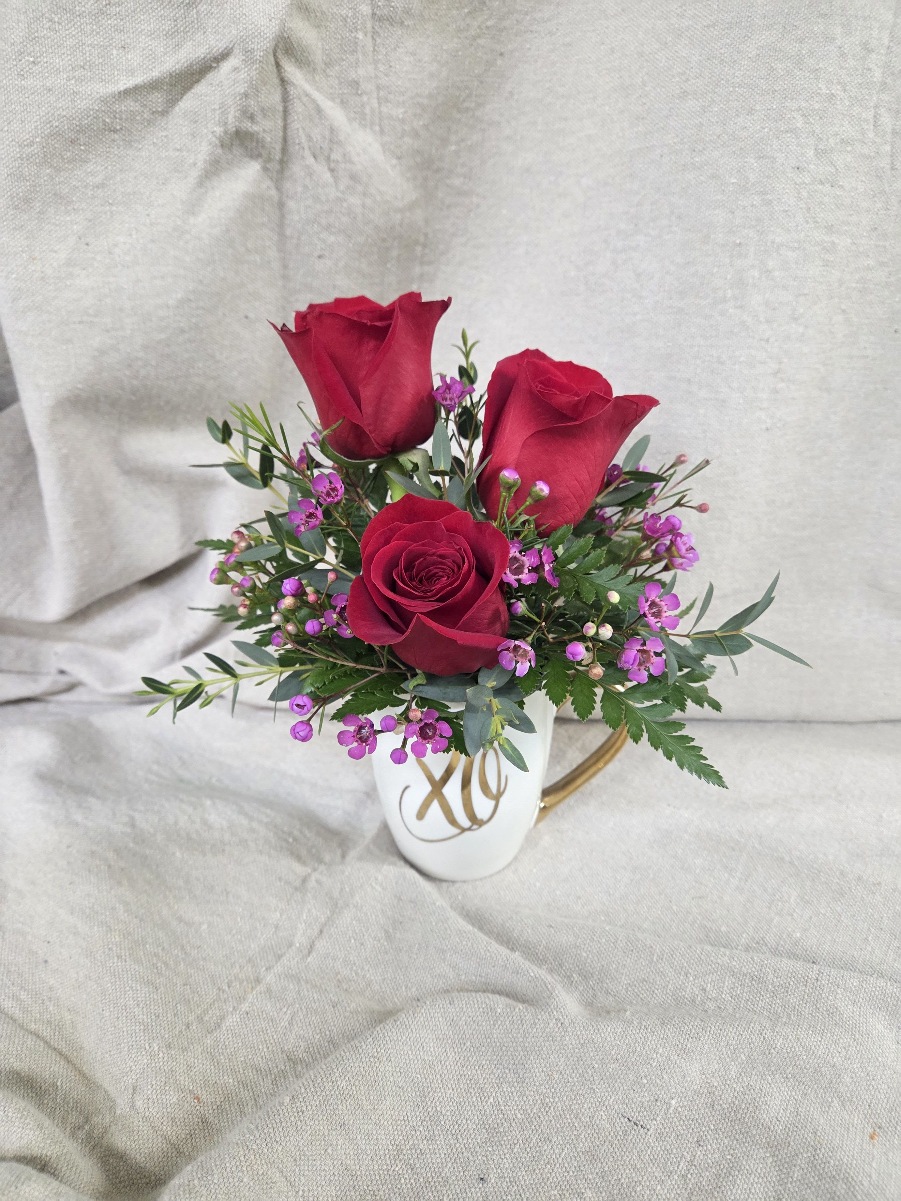 XO Crimson Roses - This elegant arrangement showcases rich red roses nestled among delicate purple waxflower and fresh, textured greenery. The deep crimson blooms create a classic expression of love, while the soft pops of purple add charm and balance. Designed in a white ceramic cup accented with gold “XO” lettering, this piece is both romantic and stylish. Perfect for anniversaries, Valentine’s Day, or any moment that calls for a heartfelt “hugs and kisses.”
