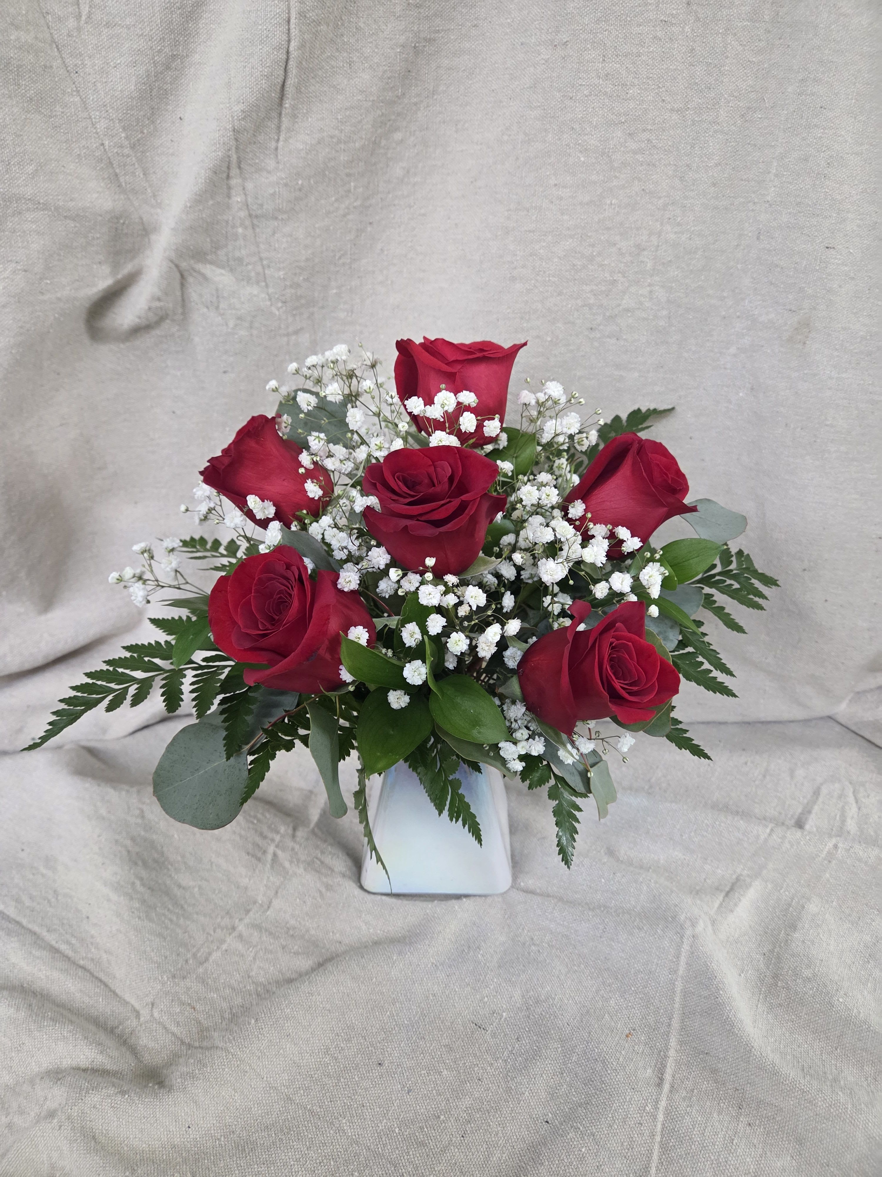 Classic Red Rose Elegance - This timeless arrangement features lush red roses beautifully paired with airy white baby’s breath and classic greenery. Designed in an iridescent vase, the rich red blooms create a striking contrast against the soft white accents and textured greens. 