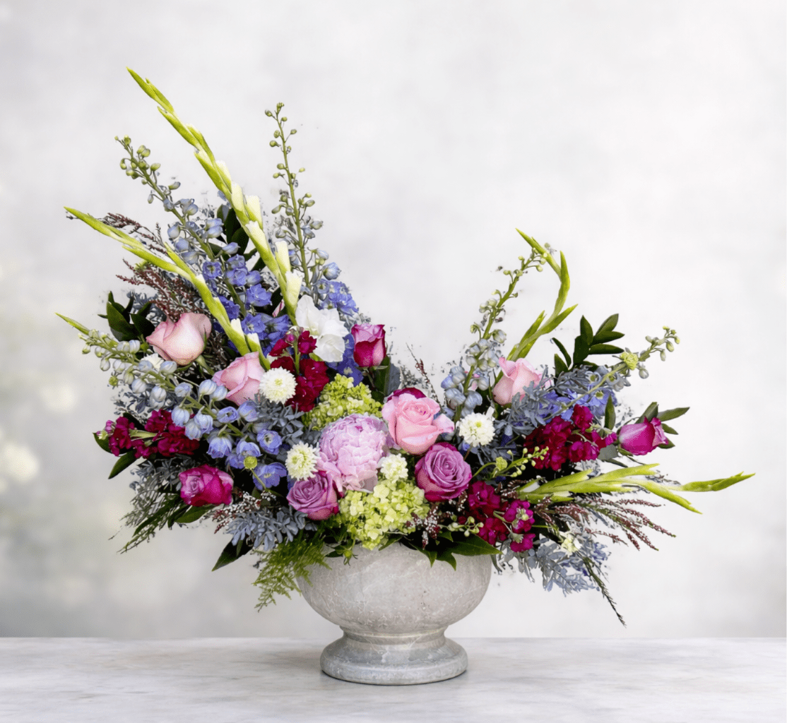 Versailles Gardens - An exquisite formal showstopper in an elegant concrete garden compote. This regal showpiece is fit for royalty with a flawless European floral style. Featuring pristine white gladiolus, blue and purple hybrid-delphinium, premium pink peony, roses, stalk, and scabiosa, framed by purple acacia, heather, and asparagus plumosa.