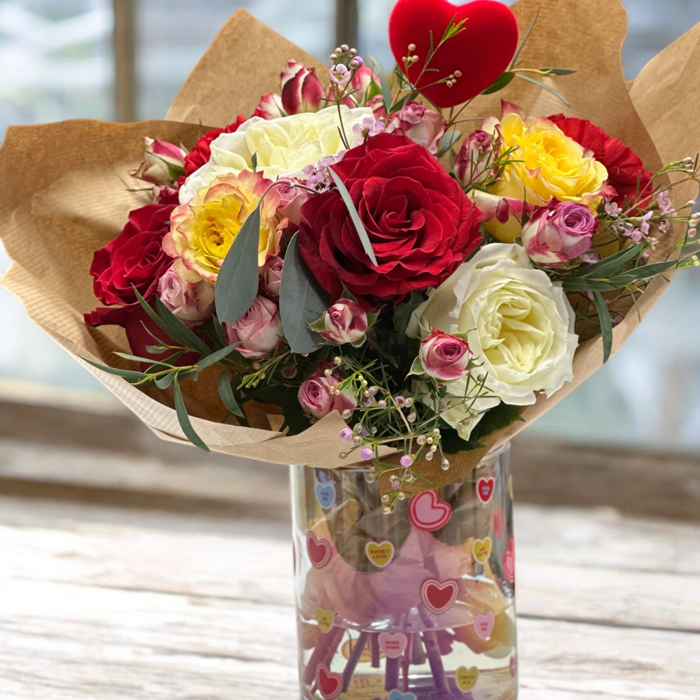 Sweet Love Bouquet - A beautiful Valentine’s Day bouquet handcrafted with love.  Designed with a romantic mix of fresh roses in red, pink, cream, and soft yellow tones, accented with delicate filler flowers and lush greenery. Wrapped in natural kraft paper and finished with a sweet heart detail, this bouquet is perfect for expressing love and appreciation.  Each bouquet is uniquely made by hand using fresh seasonal flowers.