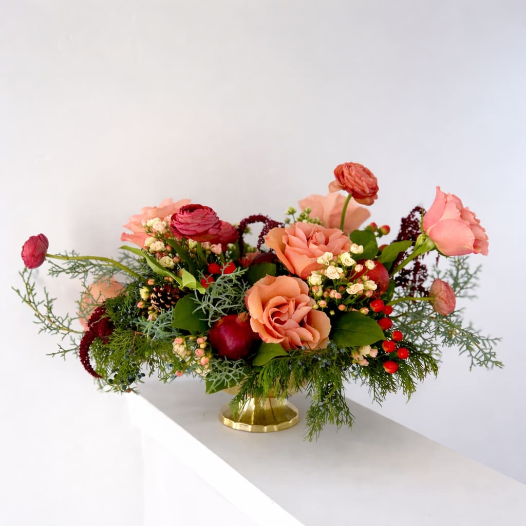 Winter Rose - A soft, romantic twist on classic holiday florals, Winter Rose brings together warm mauve roses, rich red ranunculus, winter berries, and seasonal evergreens for a centerpiece that feels both cozy and elevated. The palette leans into muted holiday tones- think plum, blush, cranberry, and cedar- creating a lush, modern arrangement perfect for winter gatherings and festive home décor.  Textural cedar, seeded greenery, and deep burgundy amaranthus add movement and depth, while soft blush accents and glossy pomegranates lend a touch of seasonal abundance. Designed in a warm gold vessel, this piece bridges timeless holiday tradition with a fresh, contemporary sensibility.