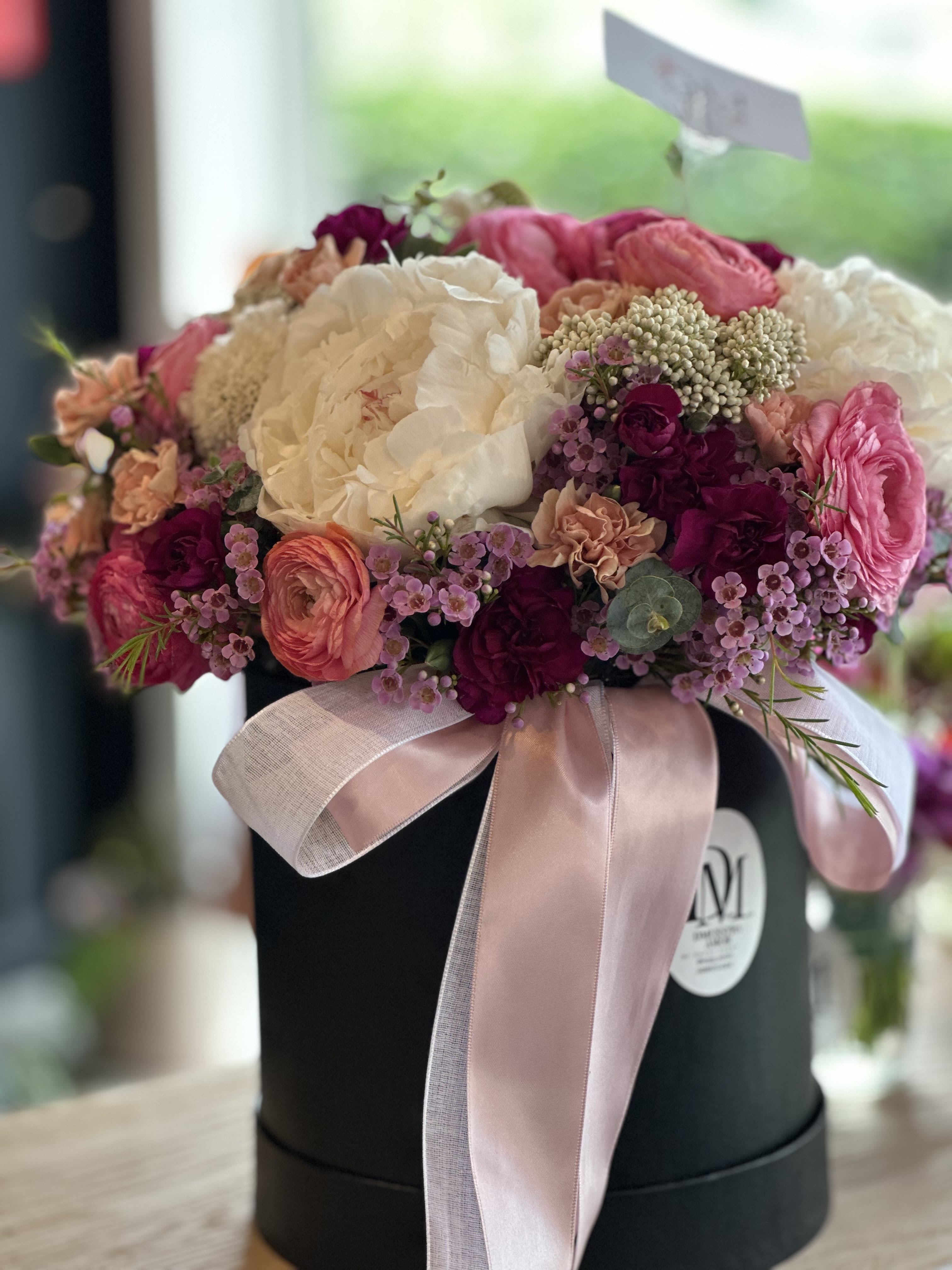 Romance in Roses – Elegant Flower Box - This exquisite floral arrangement is designed in a matte black flower box adorned with a soft pink ribbon, creating a timeless and sophisticated presentation.  The design features a harmonious blend of pink ranunculus, white peonies, deep burgundy blooms, delicate wax flowers, and soft greenery, carefully selected to convey romance, tenderness, and elegance.  Perfect for expressing love, gratitude, or celebrating special moments such as anniversaries, birthdays, Valentine’s Day, or simply surprising someone with a meaningful floral gesture.  Each arrangement is handcrafted, and while flower varieties may vary slightly due to seasonal availability, the overall style, color palette, and quality will always be preserved.   Premium flower box design 	•	Fresh, seasonal flowers 	•	Elegant presentation, ready to gift 	•	Handcrafted with care