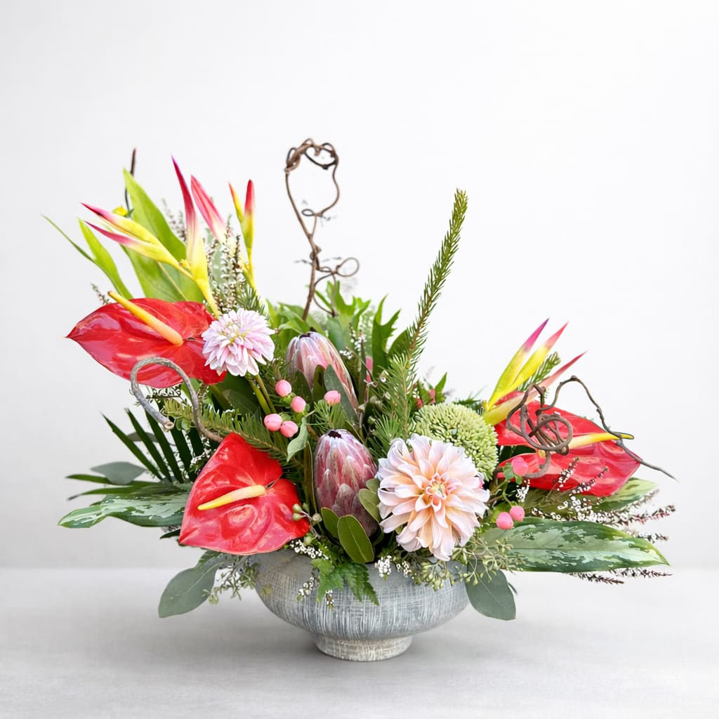 Tidalwave - Like the crest of a tropical sea, Tidalwave sweeps the eye with bold shapes and refreshing color. Striking pink anthuriums and proteas rise among soft blush dahlias, accented by cheerful yellow-and-pink heliconia and curling twigs for movement and drama. Textured greenery and berries add depth, while the arrangement’s low, ocean-toned vessel grounds it with coastal elegance. Perfect as a statement centerpiece, this design blends exotic vibrance with soft, romantic undertones — a floral tide you’ll never want to recede.