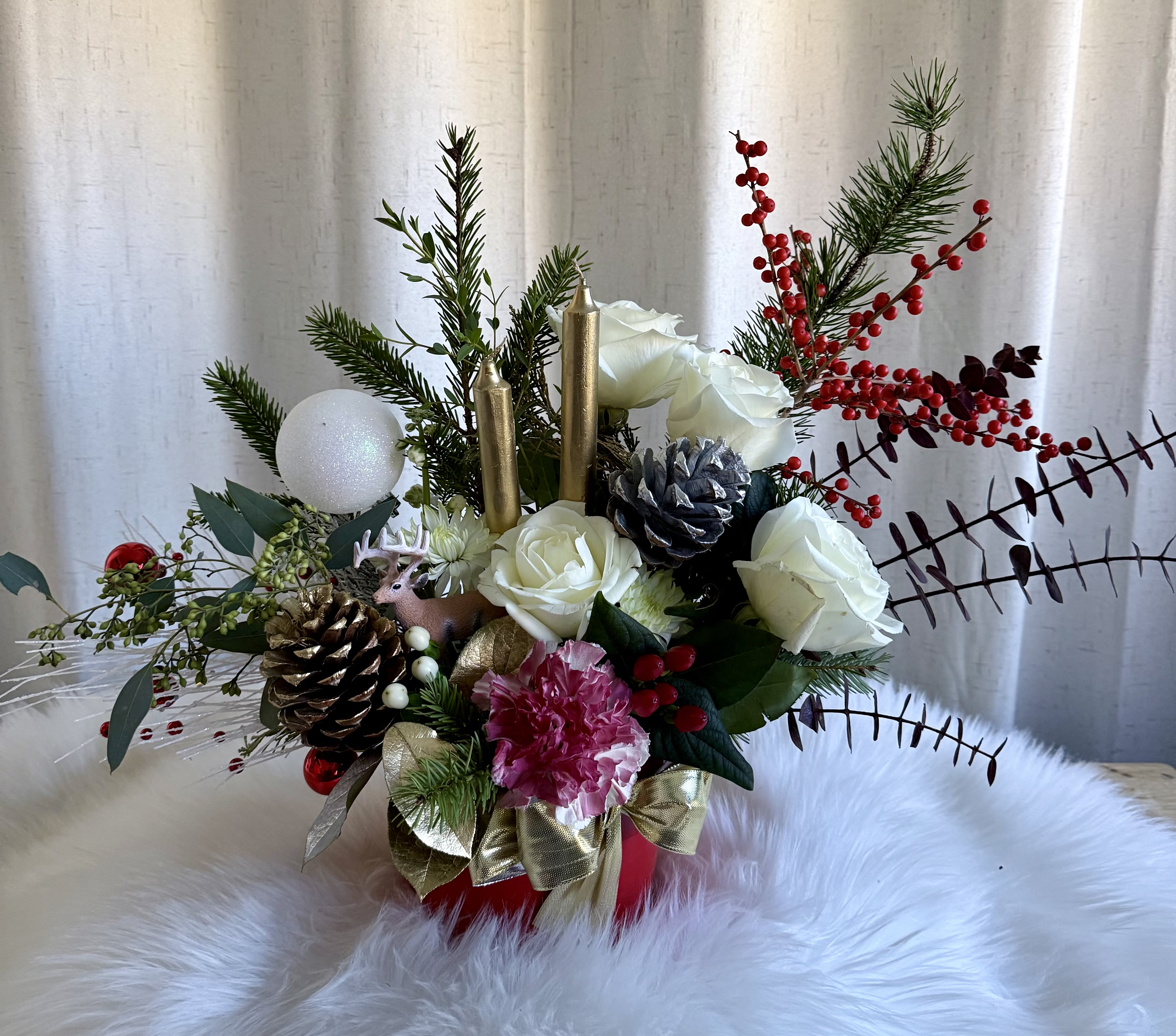  Golden Winter Candle Centerpiece - A festive holiday arrangement featuring elegant white roses, a vibrant pink carnation, gold and silver pinecones, winter greenery, red berries, and decorative candles. Accented with metallic leaves, a charming miniature reindeer, and a seasonal bow, this piece brings a warm and joyful Christmas touch to any space.