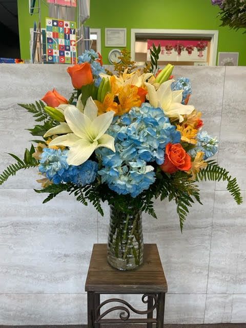 Bright Autumn Skies - A vibrant arrangement featuring **blue hydrangea**, **white lilies**, **orange roses**, and **golden yellow accent blooms**, complemented by **fresh green foliage**. The combination is bold, cheerful, and full of contrast.  