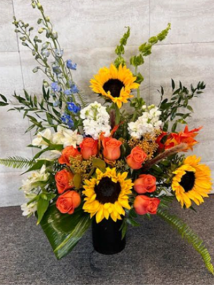 Autumn Beauty - A bold, cheerful arrangement featuring **bright sunflowers**, **orange roses**, and **white accent blooms**, complemented by **blue delphinium** and lush green foliage. The mix is vibrant, warm, and full of contrast.  