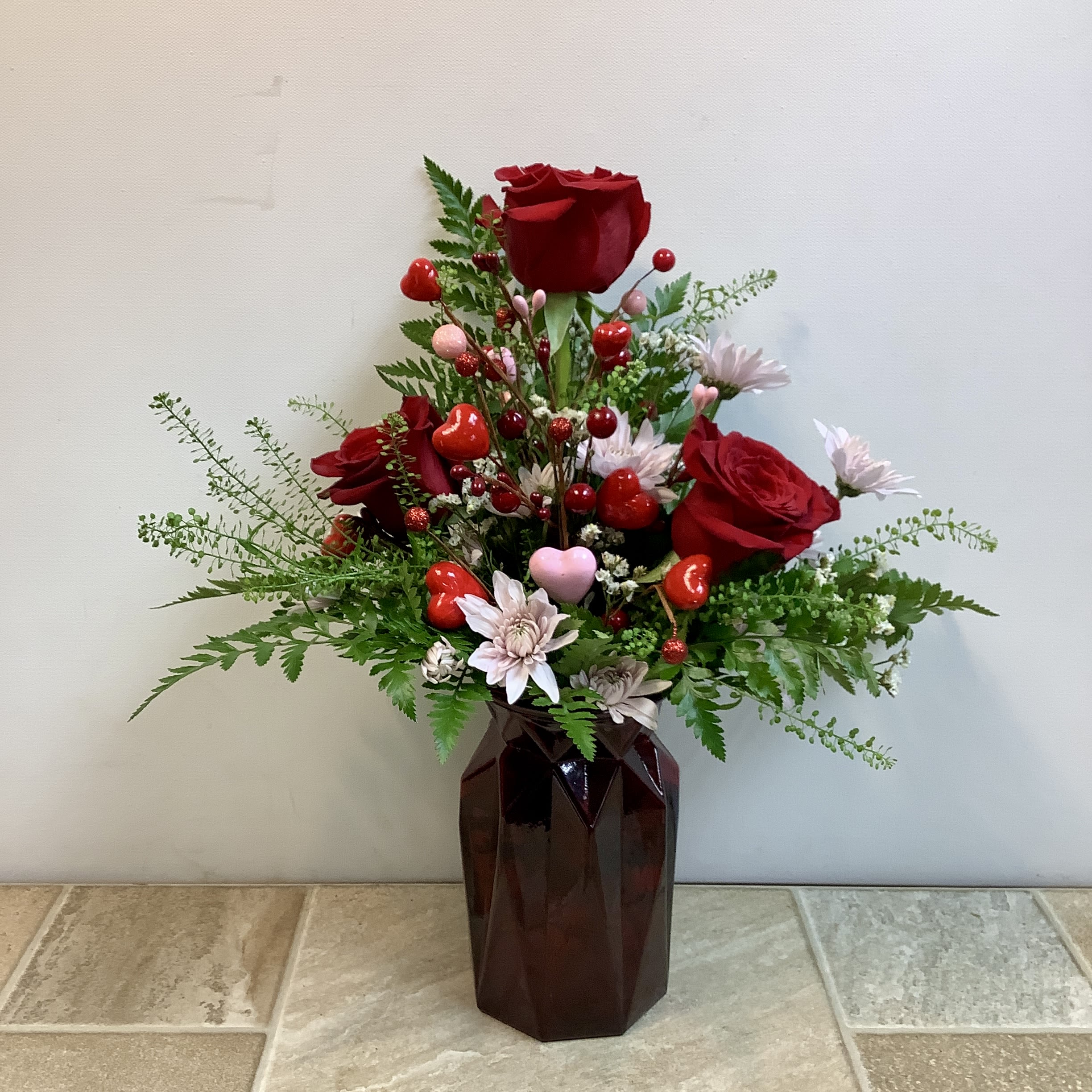 Hugs and Kisses - This romantic arrangement features rich red roses rising gracefully from a bed of lush greenery, accented with soft blush cushion mums and delicate white filler flowers. Playful red and pink heart picks are tucked throughout, adding a sweet, whimsical Valentine’s touch. Designed in a deep ruby, faceted glass vase, the bouquet balances classic elegance with heartfelt charm—perfect for expressing love, affection, or admiration. Approximate size: 13” wide x 16” tall.  Primary flowers: rose, mum Flower delivery La Crosse, Onalaska, Holmen, West Salem, La Crescent. Flower shop. Plant store. Best florist La Crosse Wisconsin area.  Seasonal and holiday floral creations. Funeral arrangements. New baby bouquets. Birthday celebration flowers. Get well arrangements. Just because gifts.