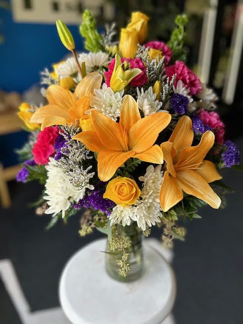 Birthday Wish - A joyful birthday arrangement bursting with color and charm, featuring radiant orange lilies, cheerful yellow roses, crisp white mums, and vibrant purple and pink accent blooms, all nestled among soft greenery. The mix of bold hues and delicate textures creates a lively, uplifting design that perfectly celebrates another year of happiness.  Beautifully crafted by Xpressions Florist, this arrangement is a bright and elegant way to say Happy Birthday and make someone’s day truly special.  