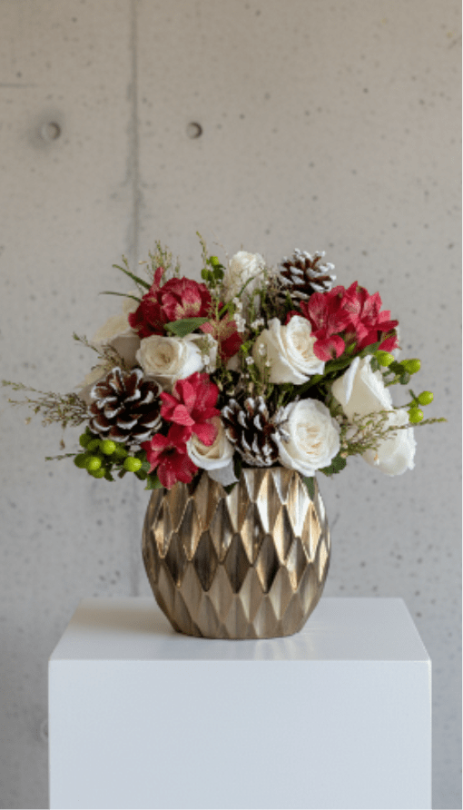 Winter Radiance - A festive winter arrangement featuring white roses, red alstroemeria, and frosted pinecones arranged in a gold geometric vase.