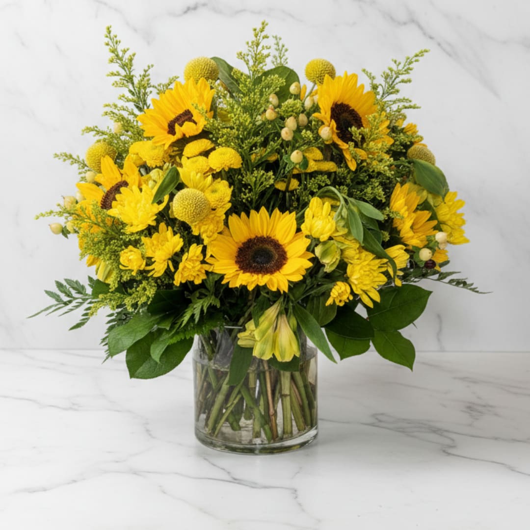 Golden Fields Beauty - Inspired by late summer meadows early fall, this lush mix of cheerful sunflowers, golden mums, hypericum berries, and accent greenery creates a vibrant and textured design. Arranged in a low cylinder vase, it’s perfect for a centerpiece or gift.