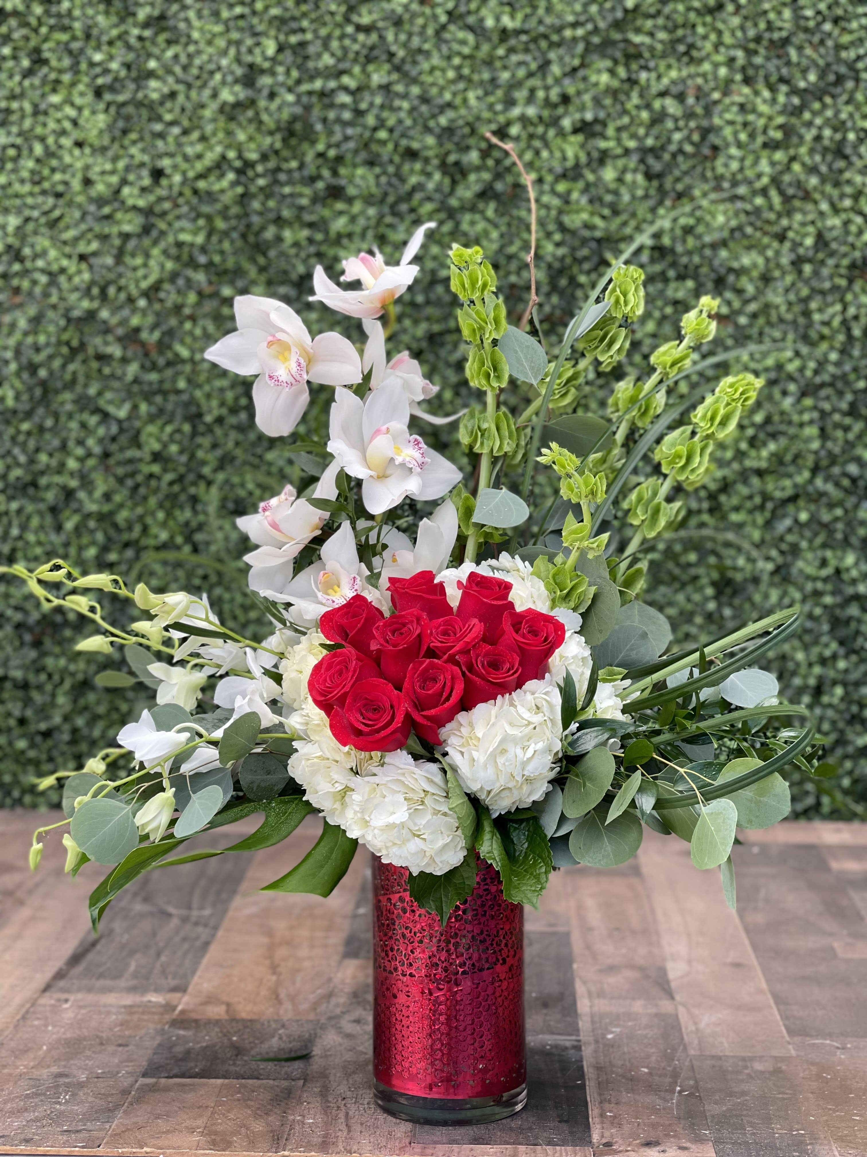 FOREVER MY VALENTINE - This modern style arrangement designed 3 sided consists of orchids, roses, hydrangeas, bells of Ireland and assorted greens.  It is designed in a clear glass cylinder with red decorative wrap inside the vase to hide the stems.