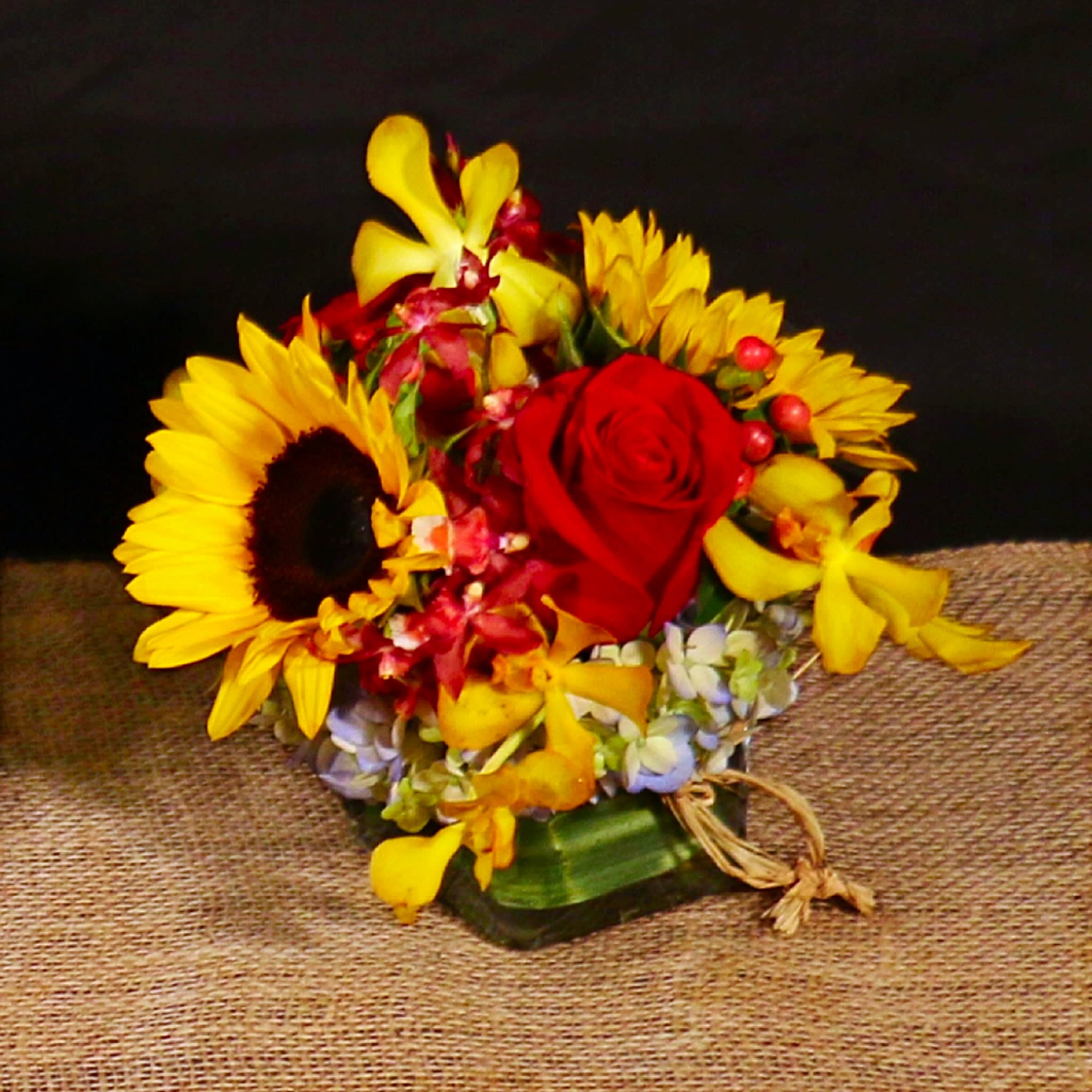 PLAYFUL MOSAIC ARRANGEMENT  - SUMMER/SPRING ARRANGEMENT EASY AND PRACTICAL IN SMALL GLASS CUBE, WITH SUNFLOWERS, HYDRANGEAS, ROSES, HYPERICUM AND ASSORTED ORCHIDS IN RED AND YELLOW.