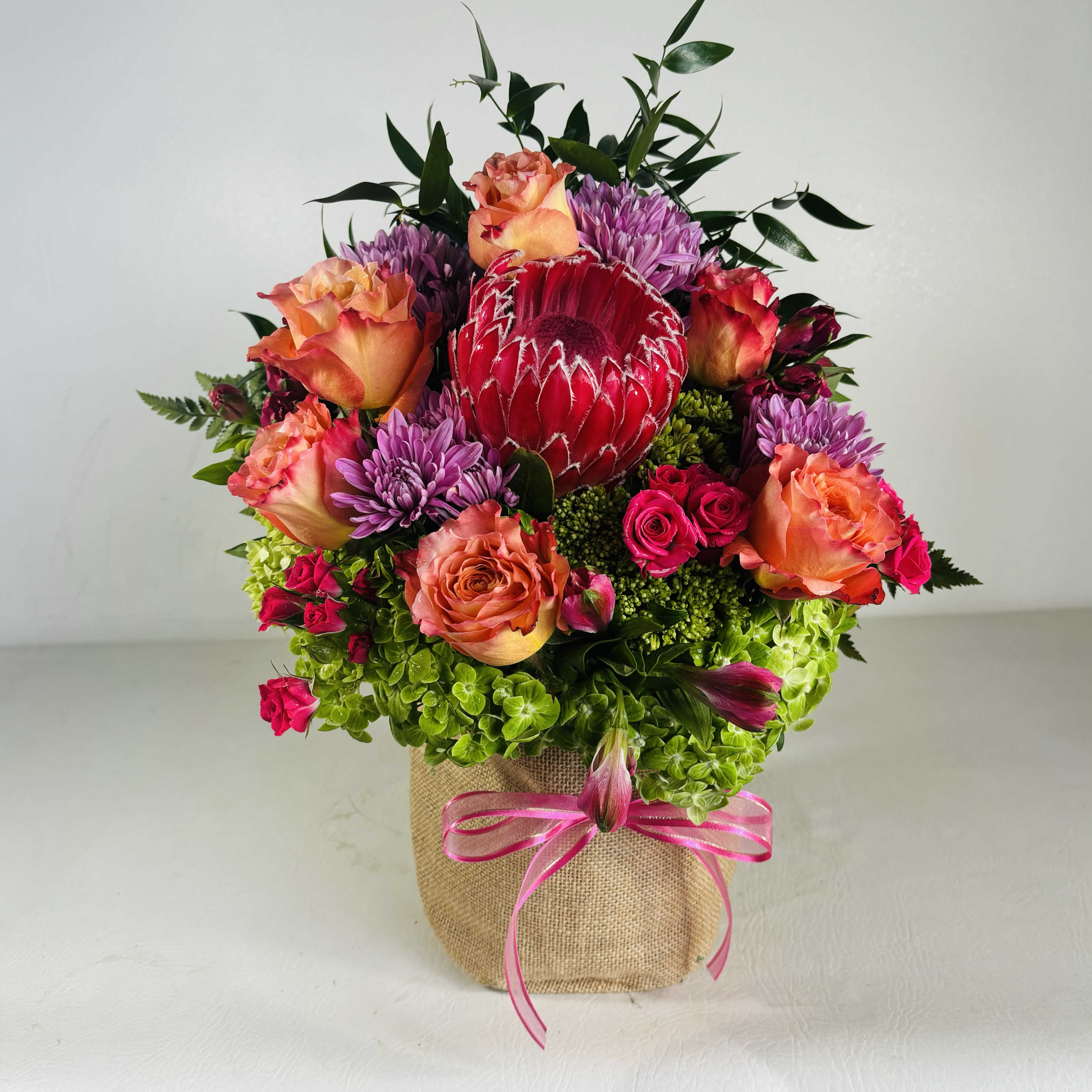 Princess Protea - A richly textured arrangement full of bold colors with a premium protea to steal the show. High magic roses, purple mums, hot pink spray roses, pink alstromerias and green hydrangeas in a rustic burlap bag.