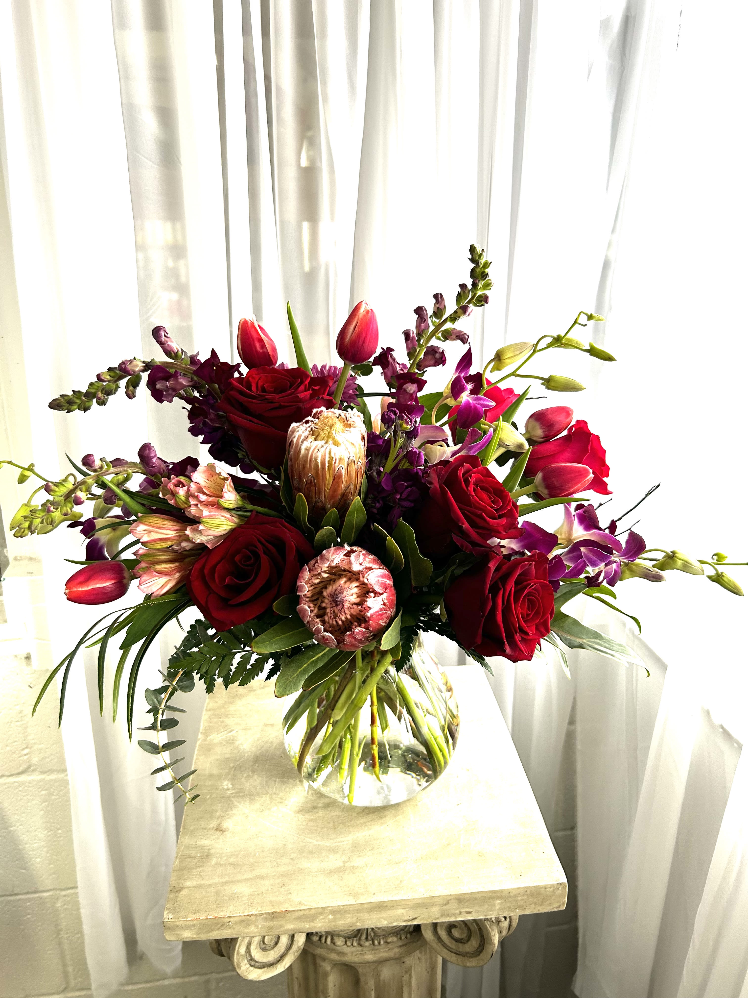 Crazy In Love - Pink mink protea, red roses, burgundy snapdragons and more are gathered in an artful display that shows how deeply you care.  Call shop if you want to know exactly what we'll be using; because our inventory constantly changes (we try to match look and feel, but probably won't be able to duplicate exactly.)
