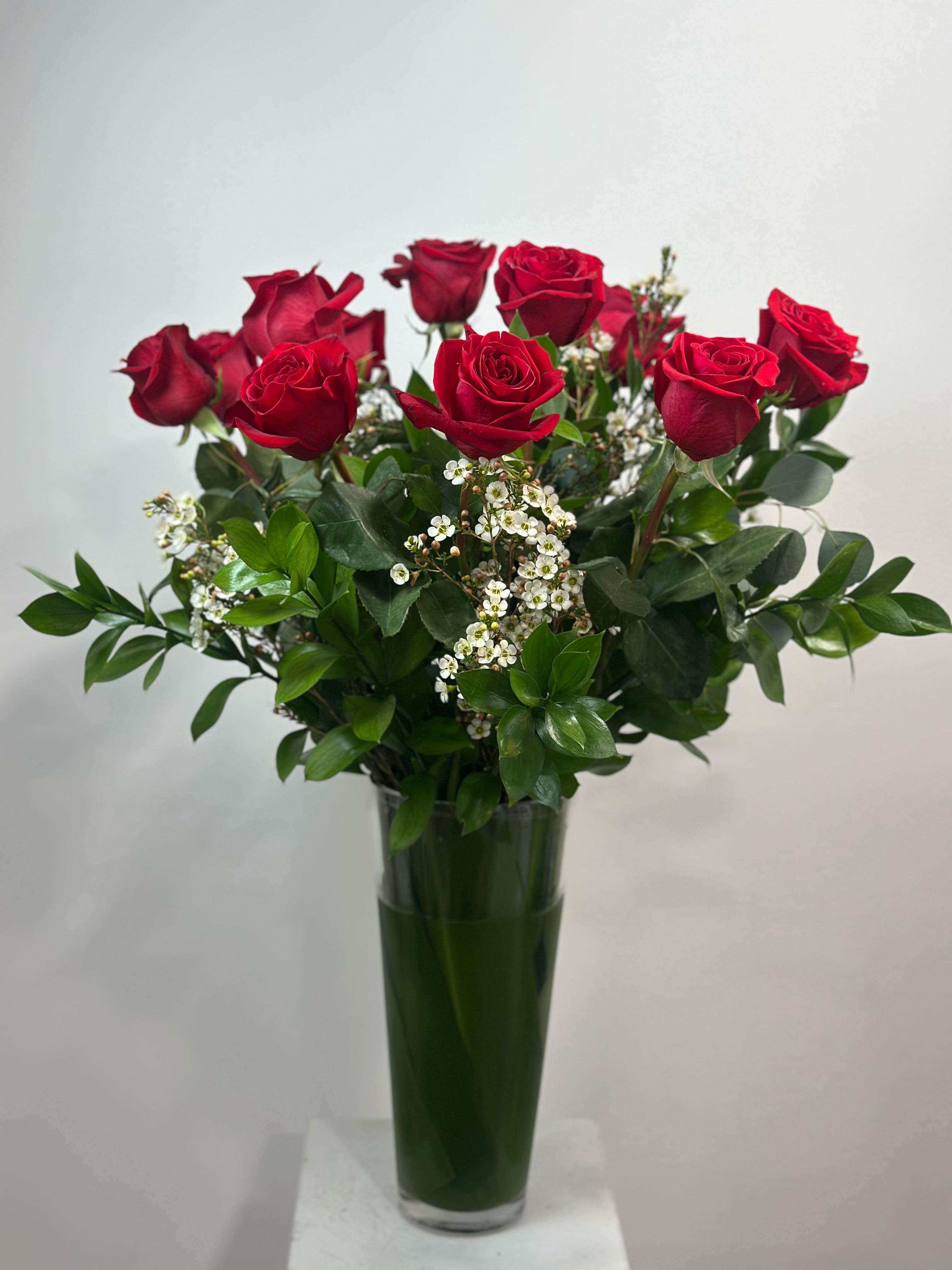 The Timeless Romance Bouquet - The Timeless Romance – Premium Red Roses in Tall Glass Vase.  Make a bold declaration of love with The Timeless Romance, a stunning arrangement designed to capture the heart. This classic bouquet features a lush gathering of premium long-stemmed red roses, universally recognized as the ultimate symbol of love and passion.  Each rose is hand-selected for its velvety texture and rich crimson hue. To enhance the natural beauty of the blooms, we accent the roses with delicate sprigs of white wax flower (or baby's breath) and frame them with deep, glossy Ruscus greenery. The bouquet is expertly arranged in a tall, clear cylinder glass vase, giving it a stately and modern silhouette that fits perfectly on a dining table or entryway console.