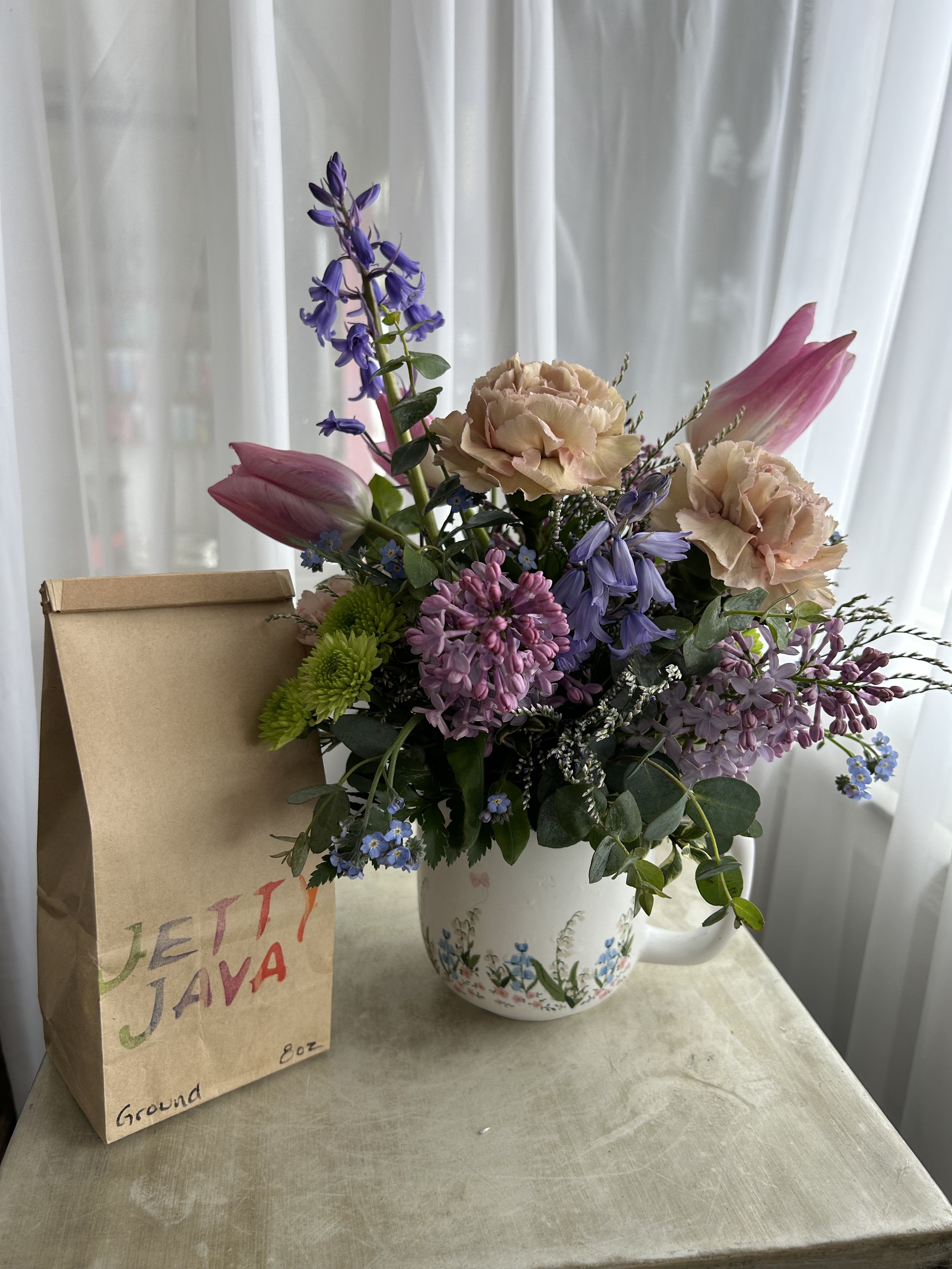 Mama Needs Her Coffee - Is your mom a coffee lover? She'll love this one. A flower arrangement inside of a coffee cup featuring the best of the season; tulips, bluebells, and lilac. Alongside a 1/2 pound bag of locally roasted Jetty Java coffee.