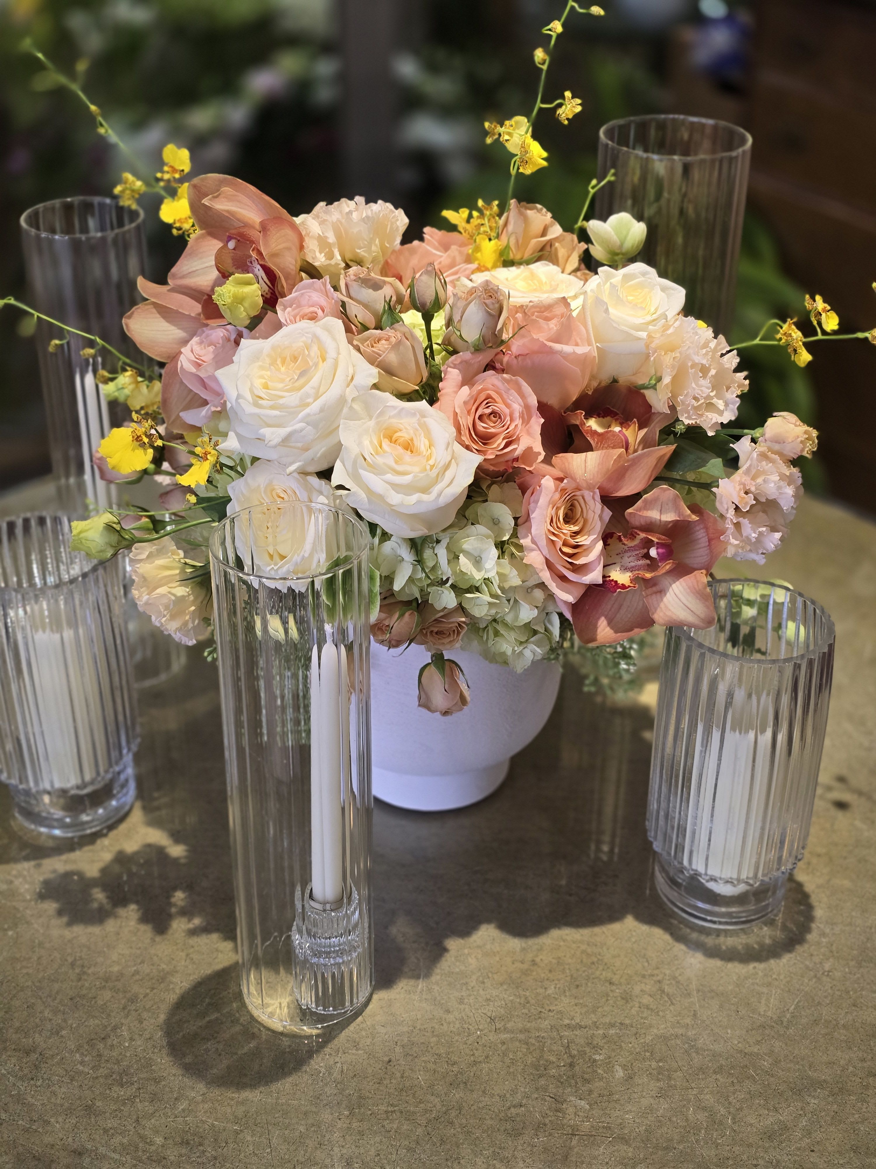 Golden Hour Embrace - Transform your table into a scene of timeless romance with the "Golden Hour Embrace." This sophisticated floral centerpiece captures the warmth of a setting sun, blending soft textures with striking architectural elements.  At the heart of this arrangement is a lush gathering of creamy white garden roses and velvety peach spray roses, offering a gradient of blush and apricot hues. The design is elevated by the exotic beauty of rust-colored Cymbidium orchids and playful sprays of yellow Oncidium (Dancing Lady) orchids, which add height and a touch of whimsy. Nestled into a base of fluffy white hydrangeas and ruffled lisianthus, the blooms sit elegantly in a modern, matte white ceramic vessel.  Perfect for wedding reception tablescapes, luxury event decor, or an intimate anniversary dinner, this arrangement is styled here with fluted glass hurricane candle holders and taper candles to create an inviting, ambient glow.