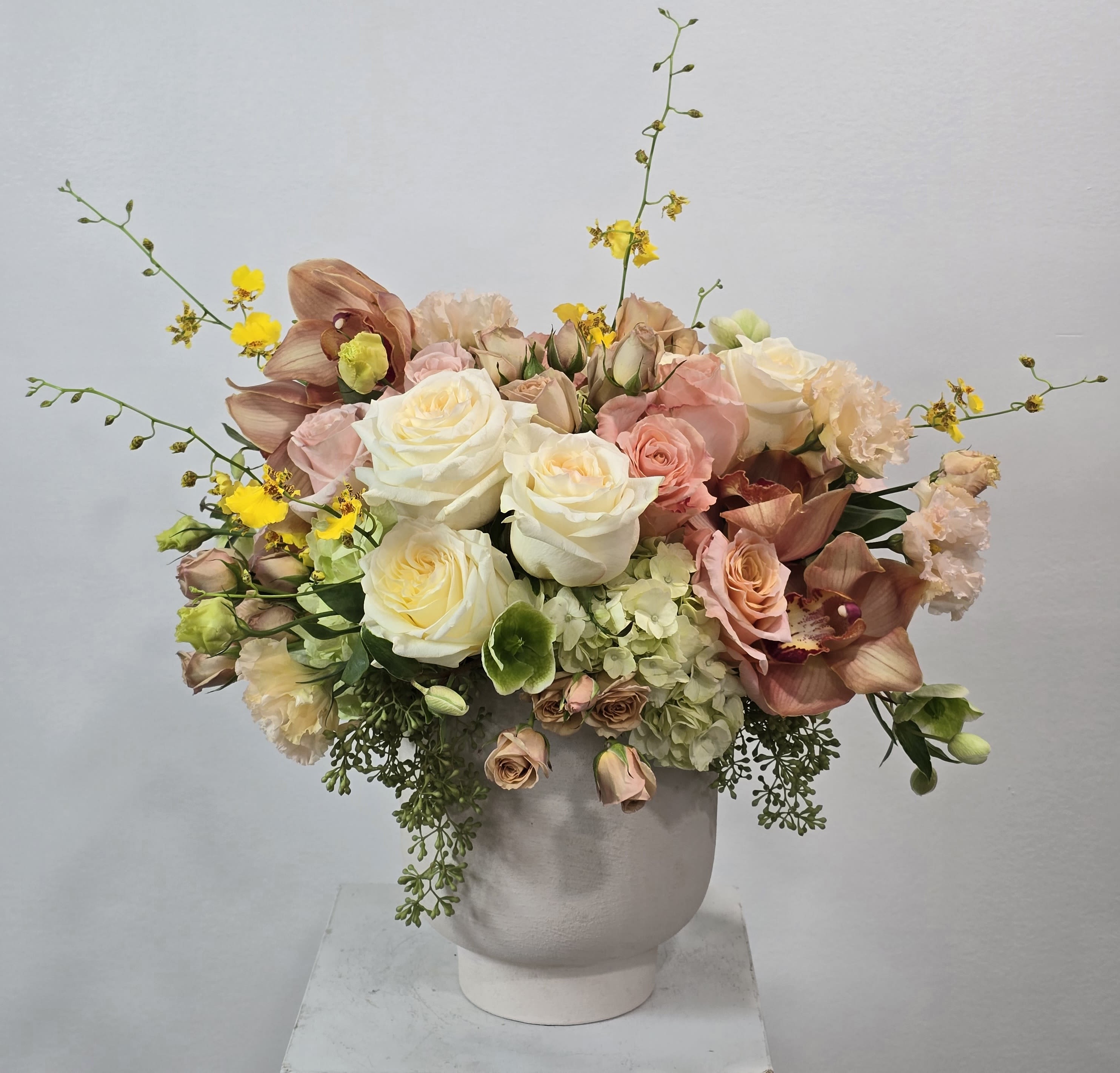 The Tuscan Sunrise - Experience texture and color in perfect harmony with The Tuscan Sunrise. This artful display bridges the gap between classic and contemporary floral design. It highlights the ruffled petals of champagne lisianthus and blush roses against the structural elegance of earth-toned orchids. Bright yellow orchid sprays add dynamic height and energy, while cascading greenery softens the edges. Presented in a minimalist, architectural white pot, this arrangement adds a touch of organic luxury to reception desks, dining tables, or modern living spaces.
