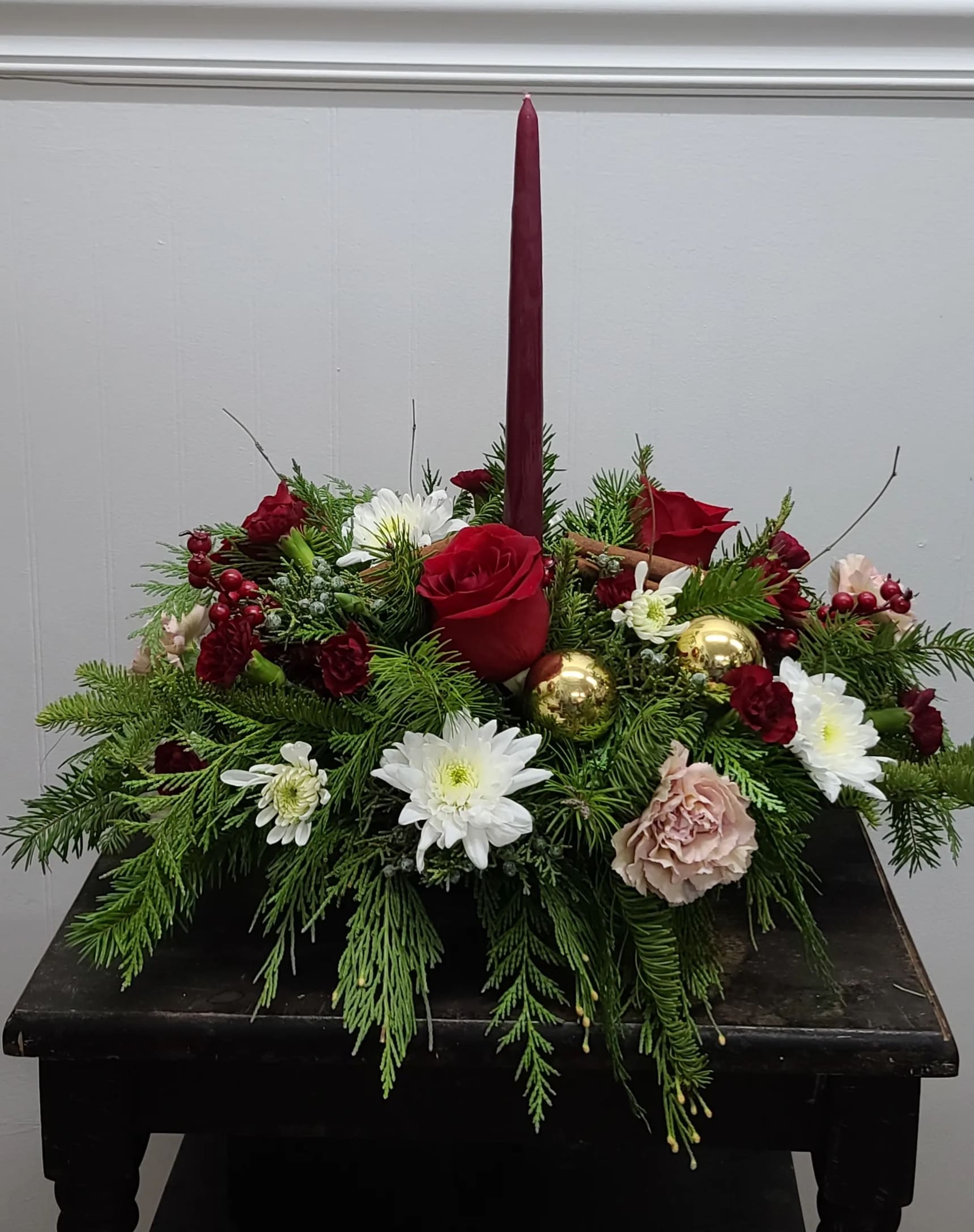 Cinnamon Spice Centerpiece - Lush mixed Christmas Greens, Cinnamon Sticks &amp; a beautiful taper candle bring this fragrant table centerpiece to life. Antique Carnations in blush, Red Velvet Roses, Burgundy mini carnations, and snow white chrysanthemums fill this arrangement with all the tradition and joy of yesterday. Send one to someone you are remembering fondly this season, today!