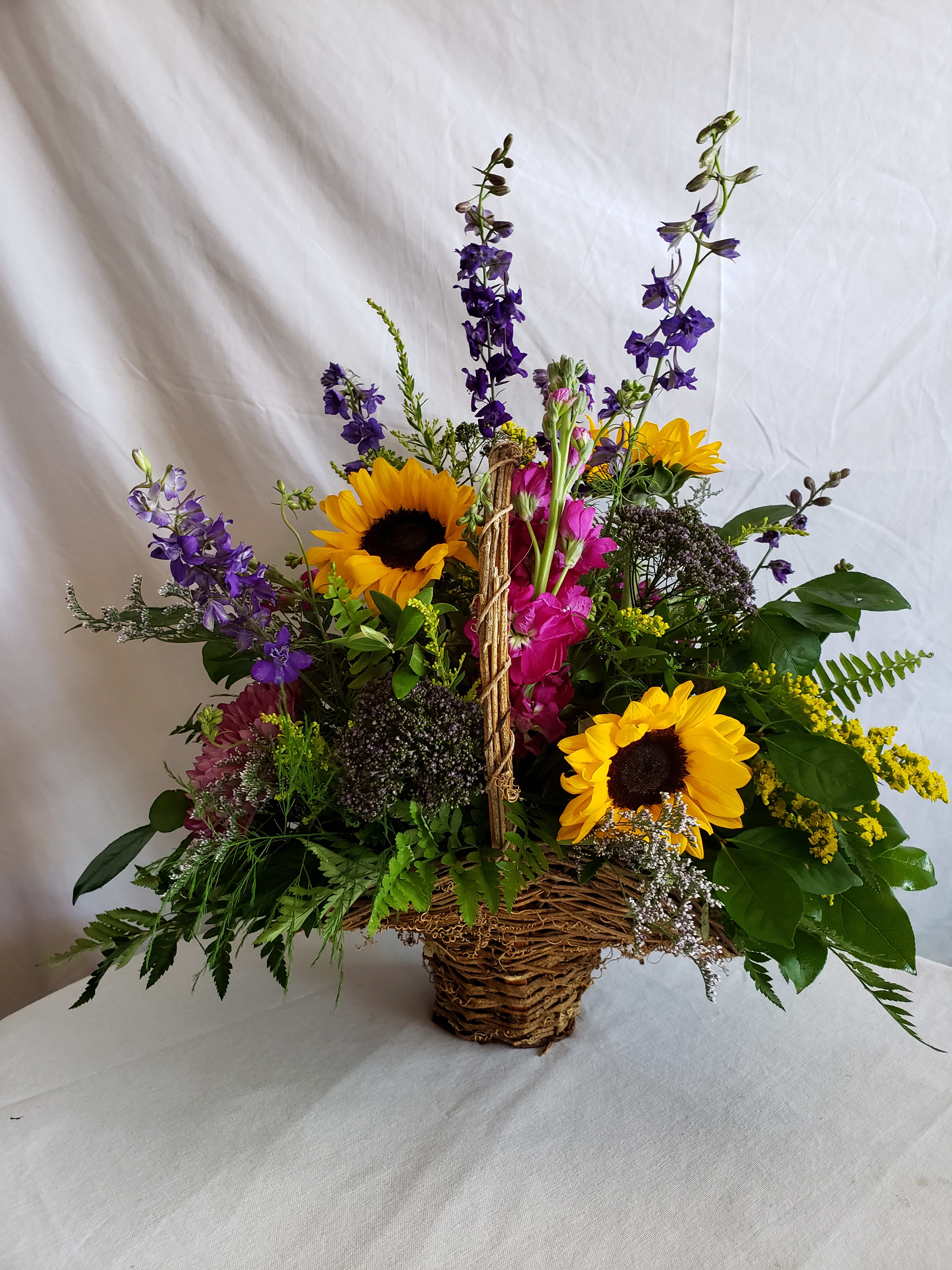 Trio of Sunflowers - This graceful grapevine basket with summers beauties in shades of Magenta, Purple and sunny yellow. Sunflowers, Larkspur, Chrysanthemums and Stock are as fragrant as they are beautiful. The easiest way to add a little sunshine to someones day. Flowers may vary by seasonal availability but overall feel will remain the same. 