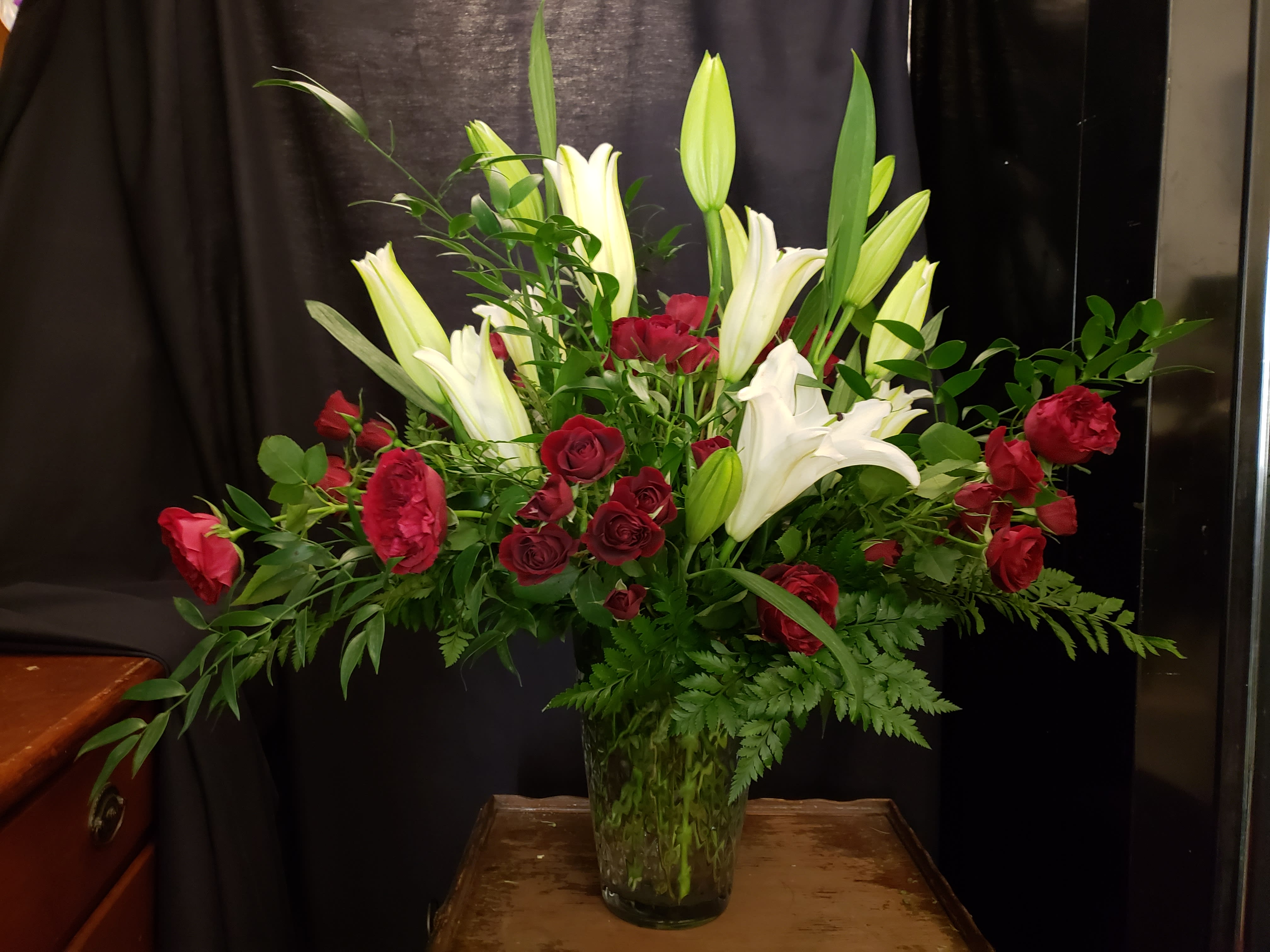 Pure Love - White lilies, Red Spray Roses and lush greenery, Pure Love is a warm embrace that lifts you up with light and joy.  Deluxe and Premium have Red Roses added to the design. (Substitutions will be made based on availability)