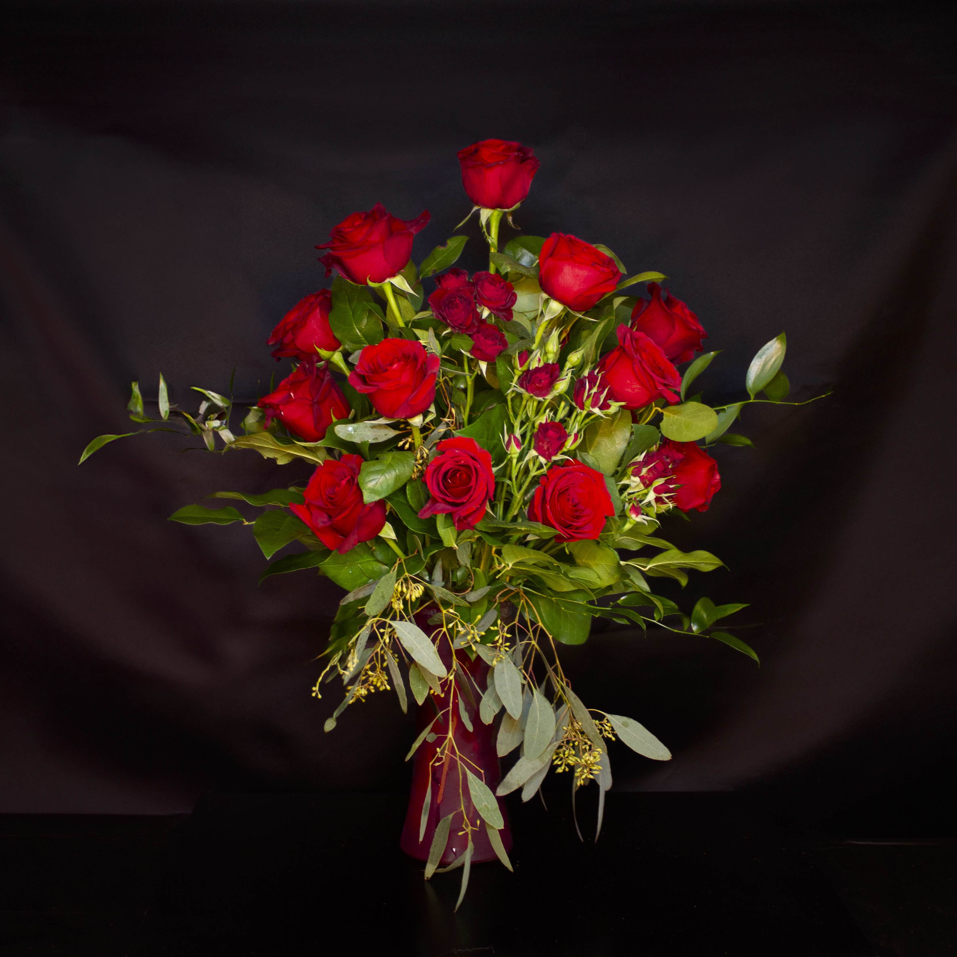'Red Romance' by Madison's Flowers -  A traditional gathering of 12 red Explorer roses with accents of scarlet red spray roses. Arranged with glossy greens and soft seeded euc, in a tall red cylinder vase. Romantic and traditional. Vase is approx. 4" x 8" .