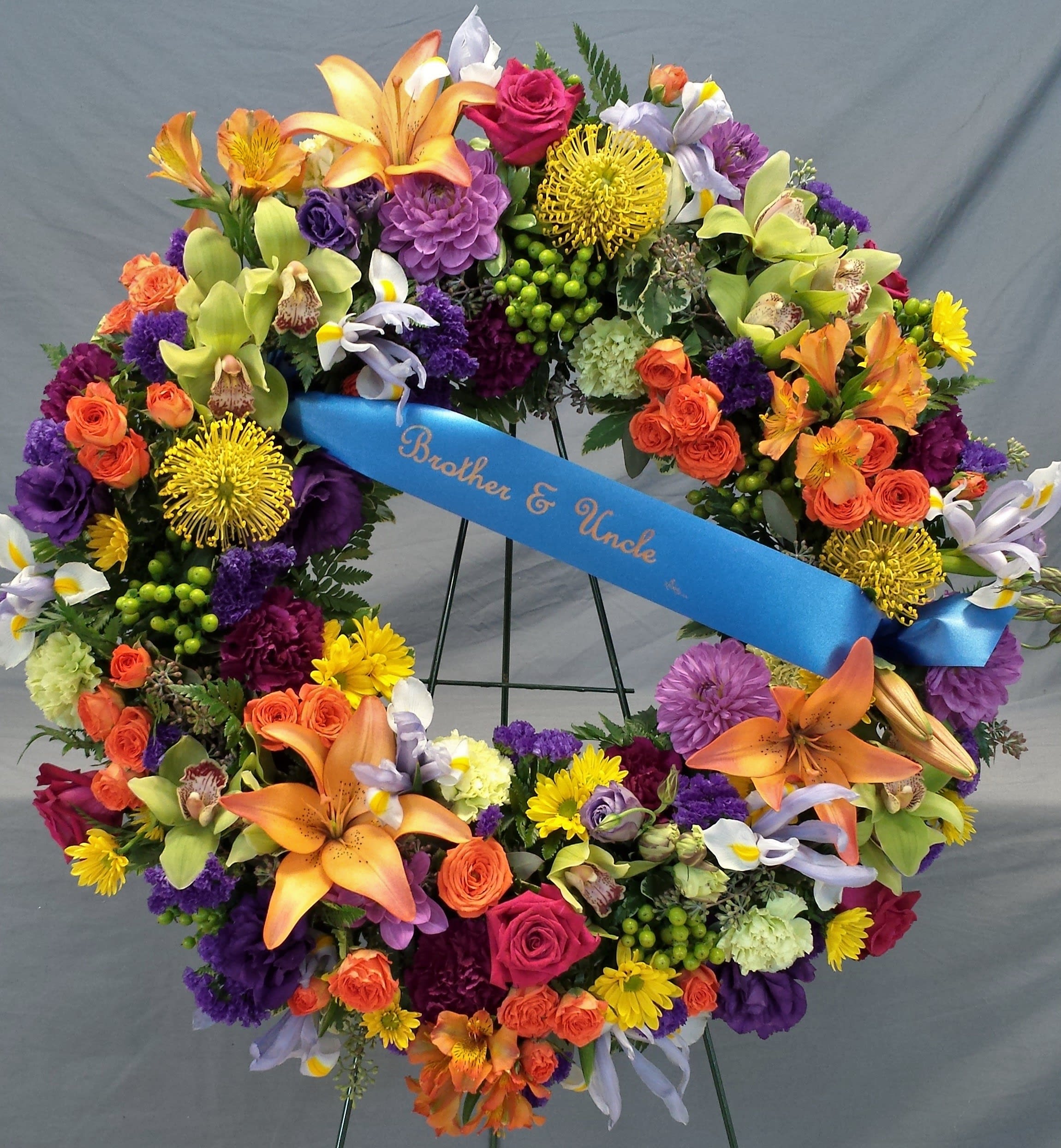 Remembrance Wreaths W13 - Bold colors dance around this wreath. Traditional blooms like iris, statice, carnations and roses are balanced with tropical orchids, protea, hypericum and more.