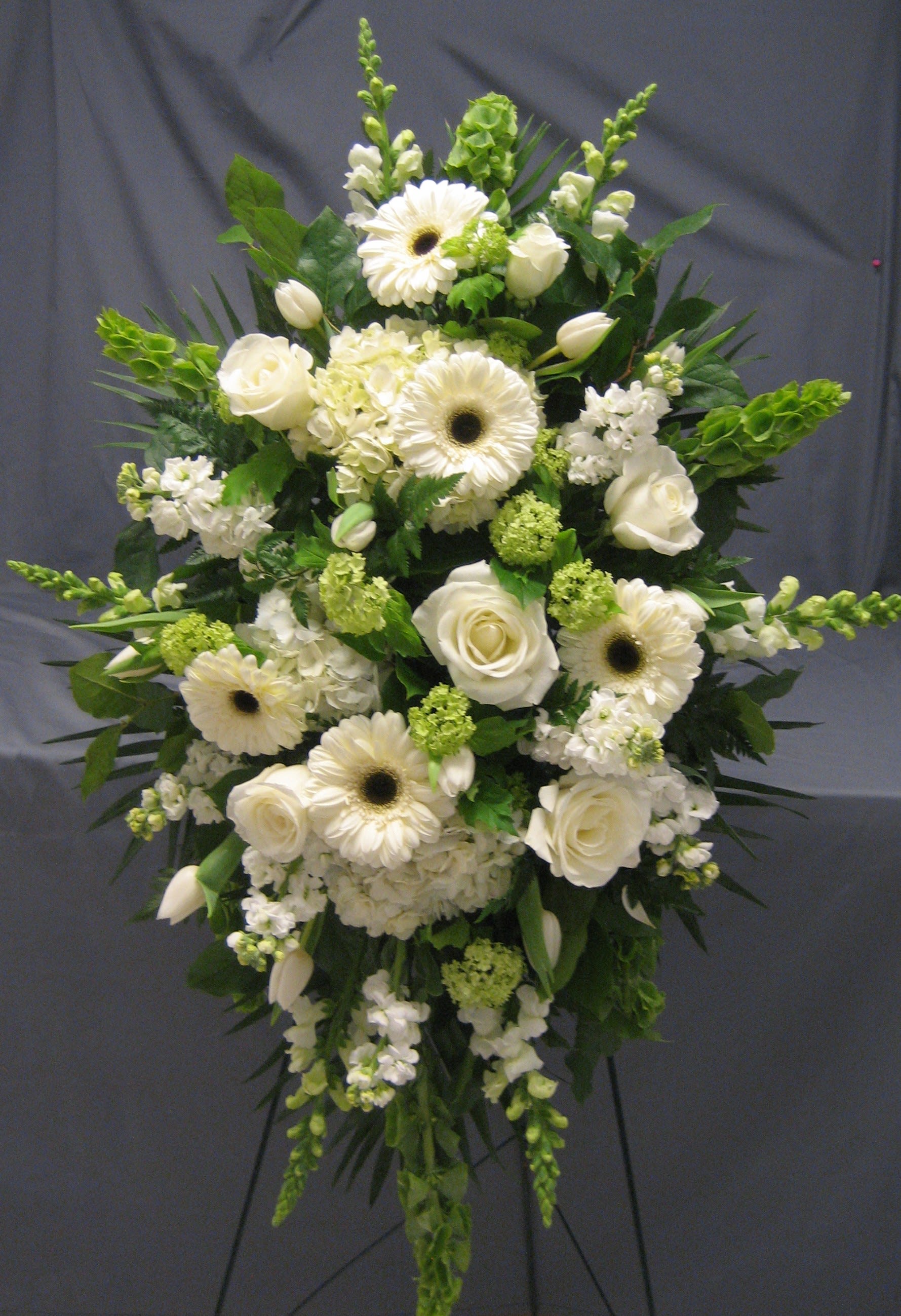 White &amp; Green Standing Spray E13 - This lush standing spray in whites and greens is a serene tribute. Bells of Ireland, snapdragons, gerber daisies, hydrangea and roses fill the elegant piece.