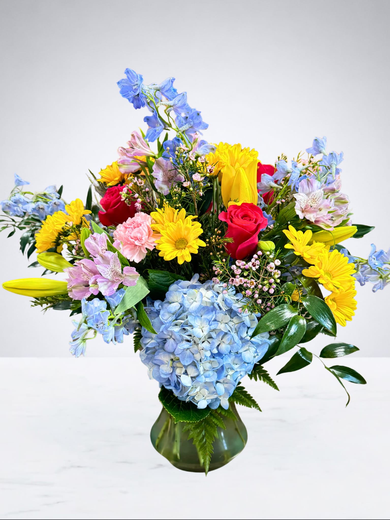 Bright &amp; Beautiful Blooms - This vibrant arrangement bursts with color and charm, featuring radiant yellow daisies, soft pink carnations, delicate blue hydrangeas, striking hot pink roses, and cheerful touches of lilies, alstroemeria, and delphinium. Arranged in a classic green vase, this design is full of life and joy, making it the perfect gift to brighten someone’s day, celebrate a special occasion, or simply bring beauty into any space.