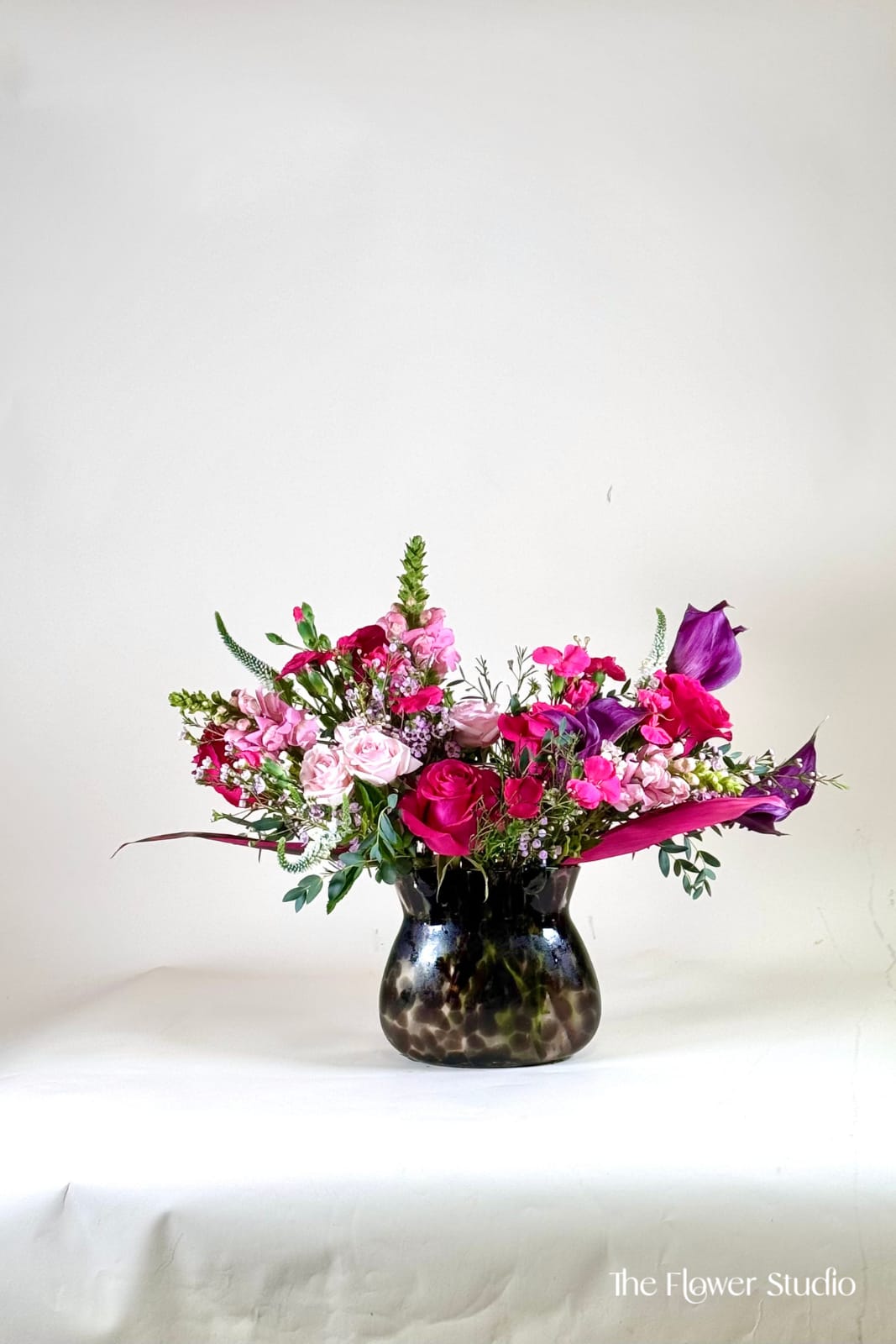 Pink Couture - A chic and fashion floral arrangement designed in a unique glass purse vase. Featuring a bold mix of vibrant pinks, soft blush hues, and rich purple accents, this design is elevated with sleek greenery for a refined, modern look. Elegant and eye-catching, this arrangement blends luxury and creativity, making it perfect for stylish gifting, special occasions, or adding a sophisticated touch to any space. Each piece is thoughtfully styled to feel exclusive and unforgettable.