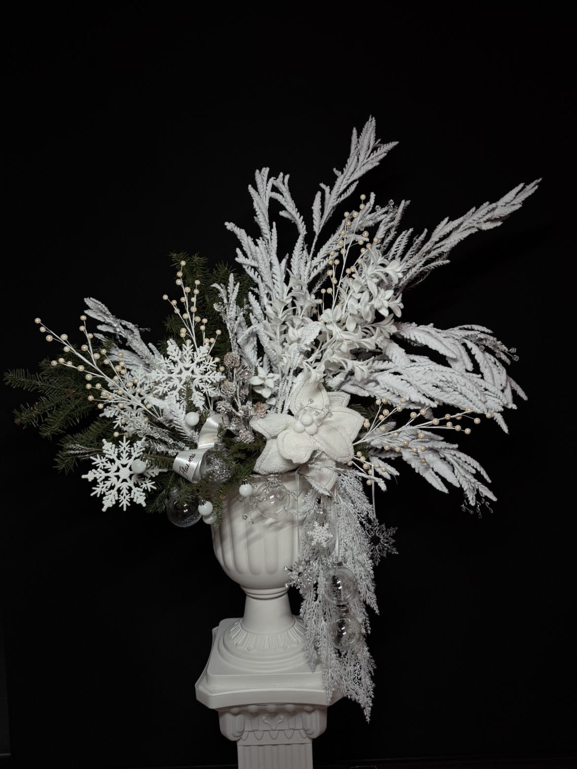 Snowfall Elegance - breathtaking winter arrangement designed to capture the magic of a quiet snowfall. Snowfall Elegance features frosted branches, soft white florals, delicate snowflake accents, pearl details, and winter greenery, all styled in a classic white pedestal vase. This statement piece radiates purity, luxury, and festive sophistication — perfect for holiday décor, winter celebrations, or elevating any interior with timeless seasonal beauty.