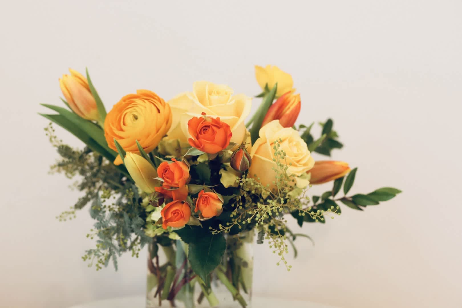 Bright and Sunny - A sweet and sunny arrangement of citrus tones, sure to bring cheer to anyone's day!  Features roses, ranunculus, and Virginia-grown tulips in a 4x4" glass cylinder.  Measures approximately 10x10".
