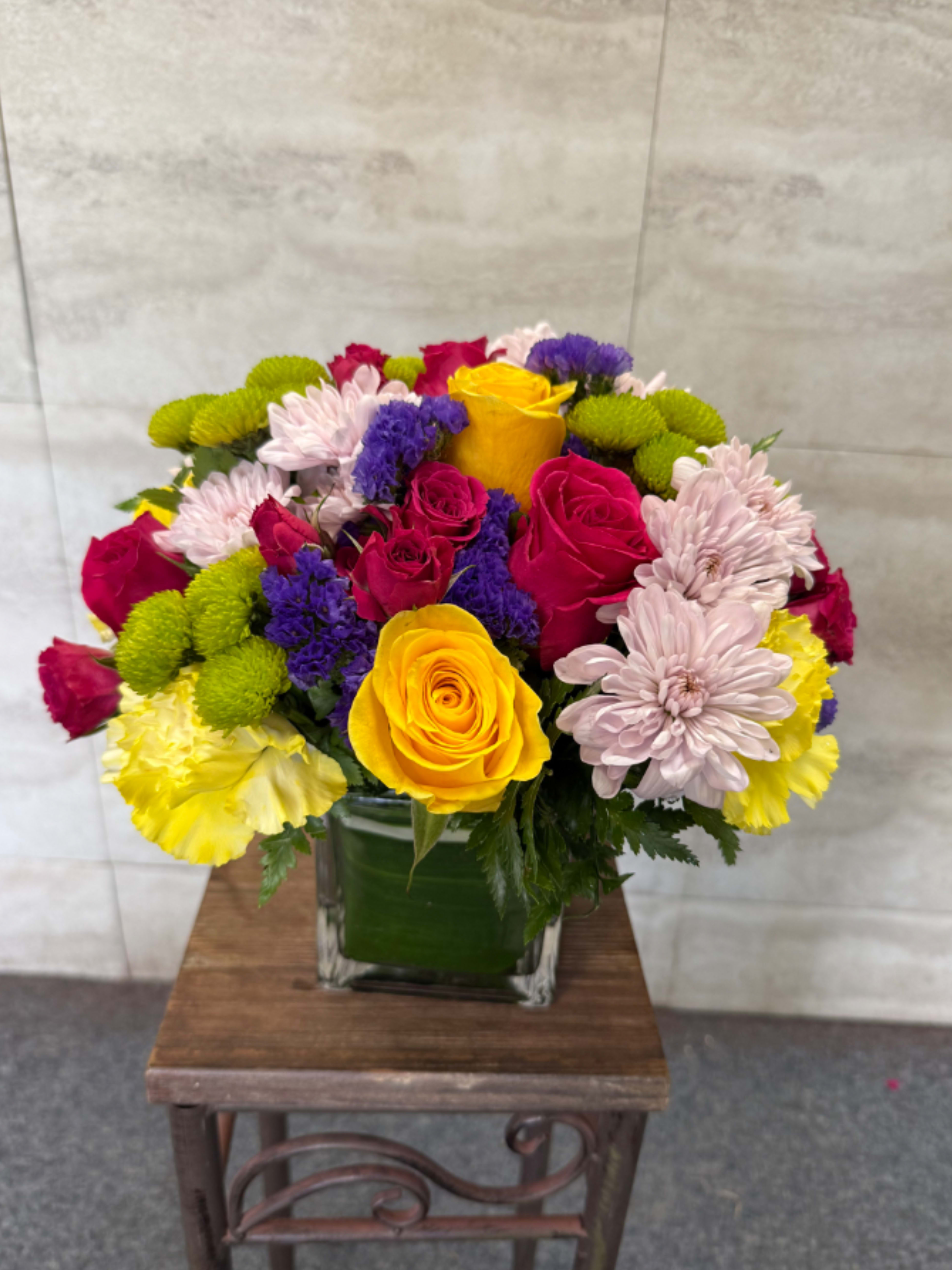 Bright Remembrance - A graceful and uplifting arrangement featuring vibrant yellow roses, rich pink roses, soft lavender chrysanthemums, green button mums, and accents of purple statice. Designed in a clear glass cube, this colorful bouquet offers warmth, comfort, and a gentle reminder of cherished memories during times of sympathy and reflection. 