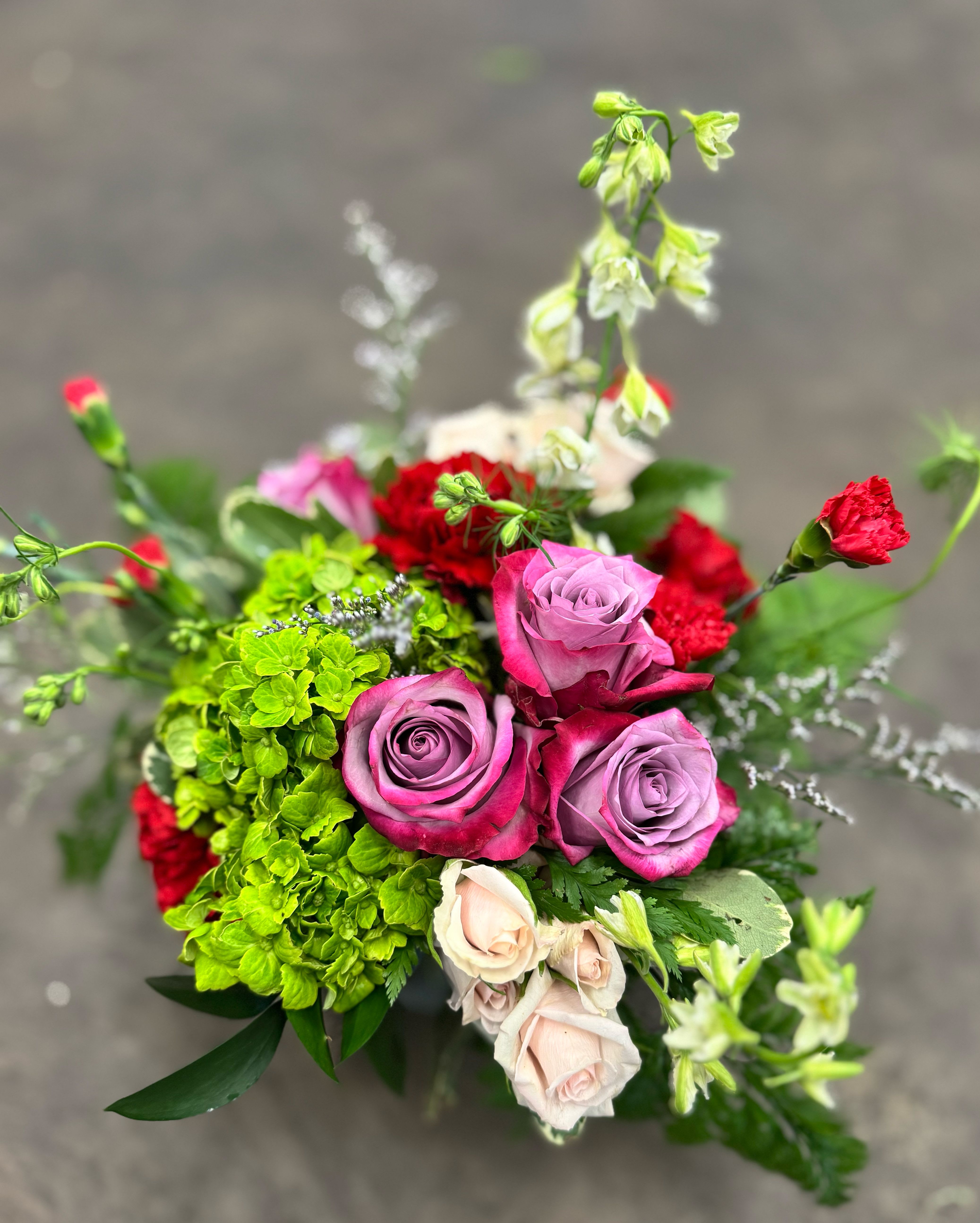 Rosewood - A lush blend of lavender roses, rich red carnations, green hydrangea, blush spray roses, white Larkspur and delicate filler blooms, thoughtfully arranged in a glass vase with fresh greens. Bold yet balanced, this design is perfect for birthdays, anniversaries, or sending something beautiful “just because.”