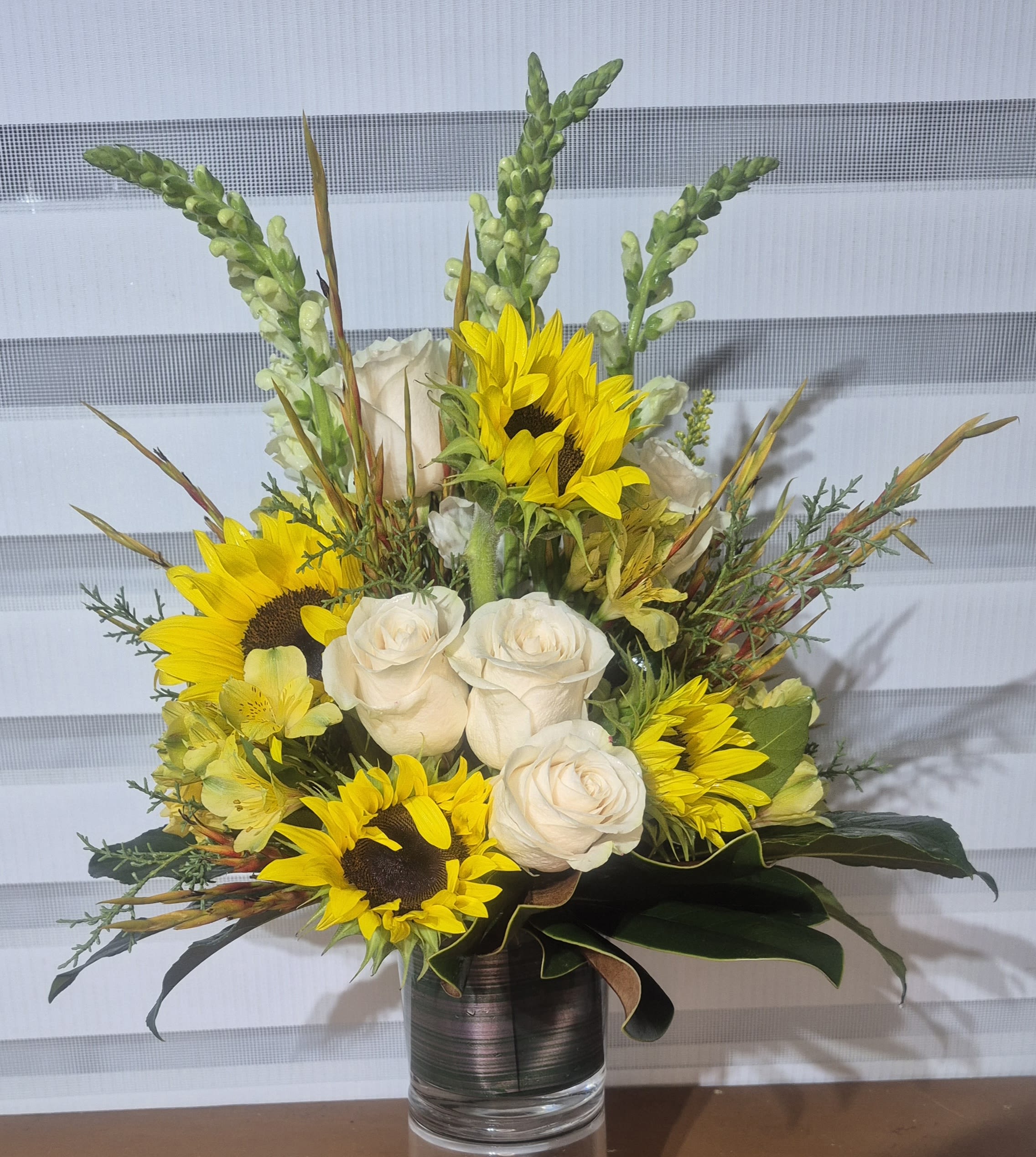 YELLOW SUNRISE - AN ALL AROUND ARRANGEMENT IN A 5X5 LEAF WRAPPED GLASS VASE. ARRANGEMENT CONSISTS OF WHITE ROSES, WHITE SNAPDROGONS, YELLOW ALSTROMERIAS, YELLOW SUNFLOWERS AND EXOTIC GREENS AND FILLER.ELEGANCE.