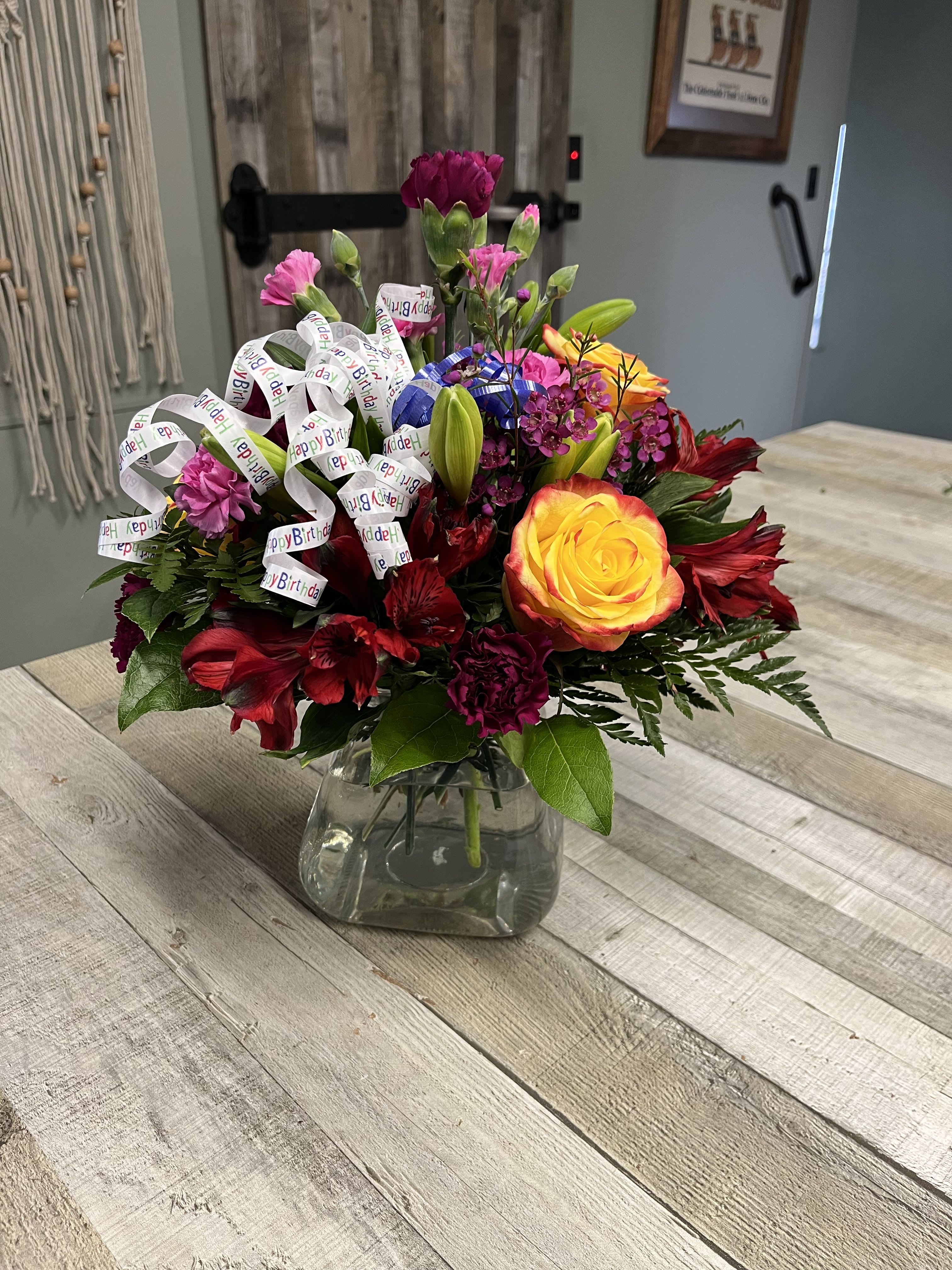 The Birthday Cheer Bouquet - Bright  blooms that are ready to get your recipient's special day started this flower bouquet is bright happy and ready to celebrate. Roses and  other vibrant flowers  surrounded by purple Peruvian Lilies,  mini carnations and filler and an assortment of lush greens create this unforgettable birthday bouquet. We will choose the best selection of flowers and colors for you. Arrangement is in a low glass cube vase. 