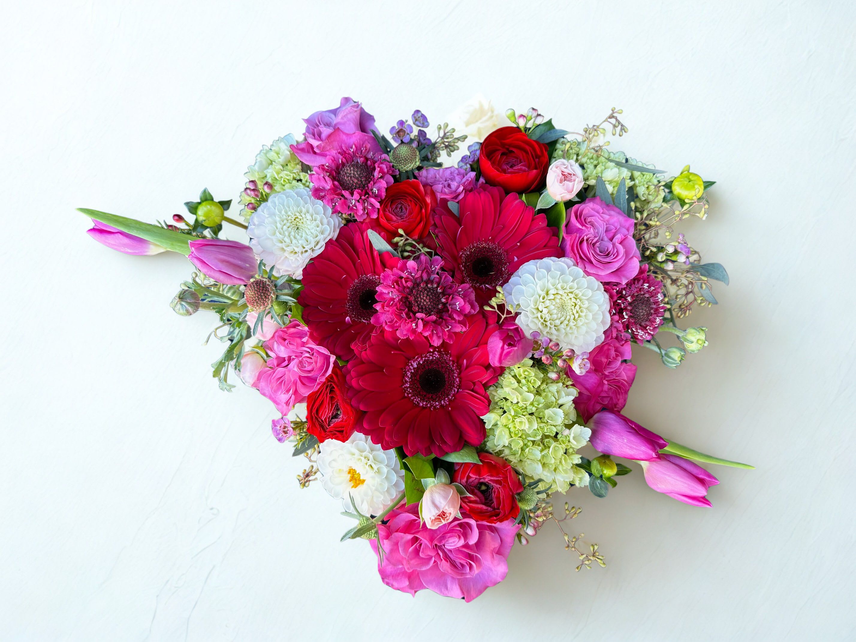 Heart Burst - Lush, colorful, and full of joyful energy, this garden-inspired arrangement is a stunning blend of bold reds, vibrant pinks, and fresh greens. Featuring striking red gerbera daisies, romantic roses, soft pink tulips, delicate white blooms, and textured green hydrangea, each flower is thoughtfully arranged for depth and movement. Designed in a wood heart box, this a the top view picture. 