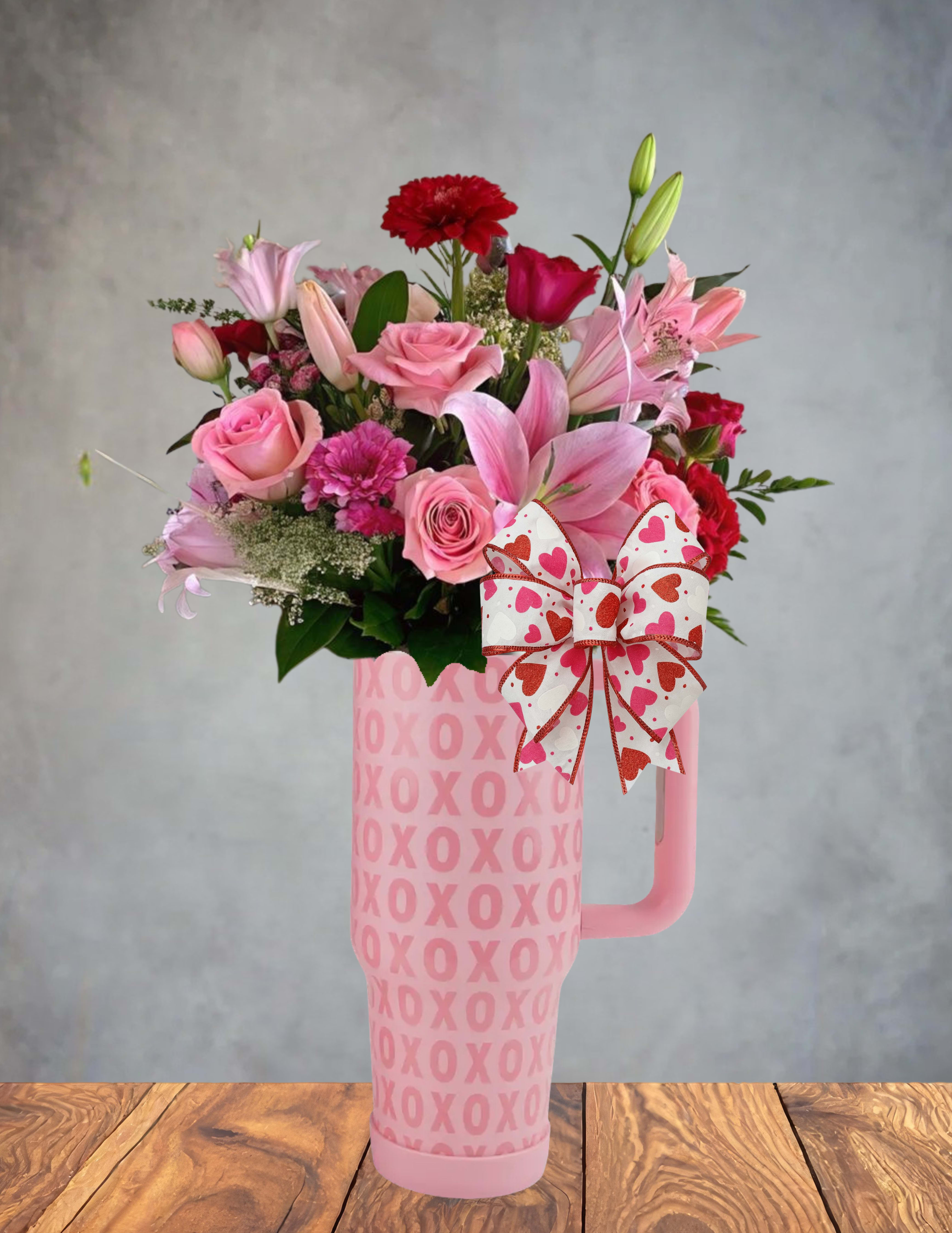 XO Overflow - A celebration of love that can’t help but spill over. XO Overflow bursts from its pink “hugs &amp; kisses” tumbler with a lush mix of roses, lilies, carnations, and soft seasonal blooms in shades of blush, berry, and whisper‑white. Every petal feels like a little love note, gathered together in a bouquet that’s full, flirty, and overflowing with affection.  Finished with a charming heart‑patterned bow, this arrangement is perfect for anyone who deserves a little extra sweetness—Valentine’s Day, anniversaries, or those everyday moments when “thinking of you” just isn’t enough.