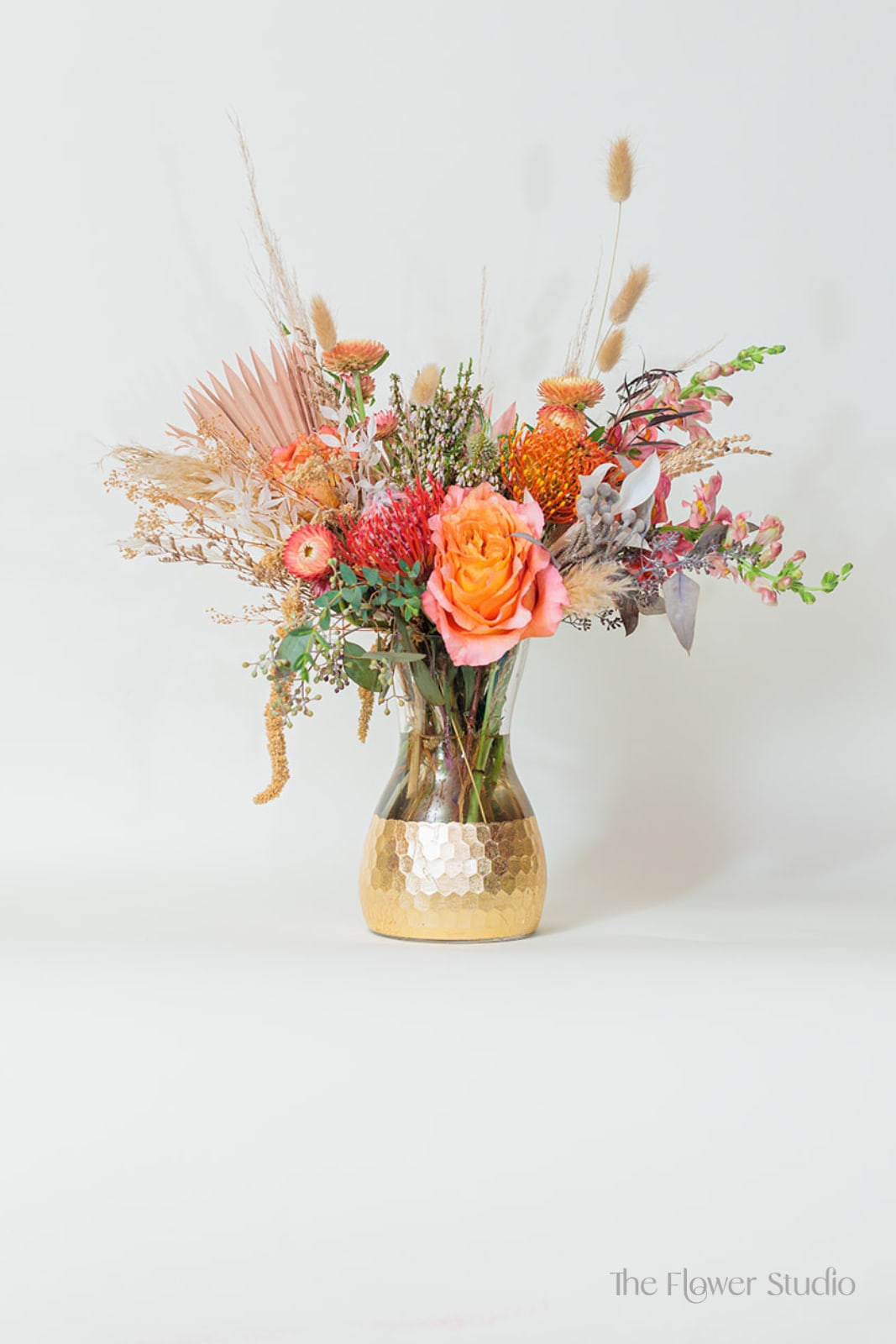 Boho-ho - A stunning celebration of warm hues and natural textures, this arrangement features vibrant orange roses, symbolizing passion and energy, alongside a striking pincushion that adds an intriguing focal point. Displayed in a golden vase, this arrangement exudes warmth, grace, and everlasting beauty making it a perfect way to honor a loved one or celebrate a special occasion. Approx. H16” x W15” 