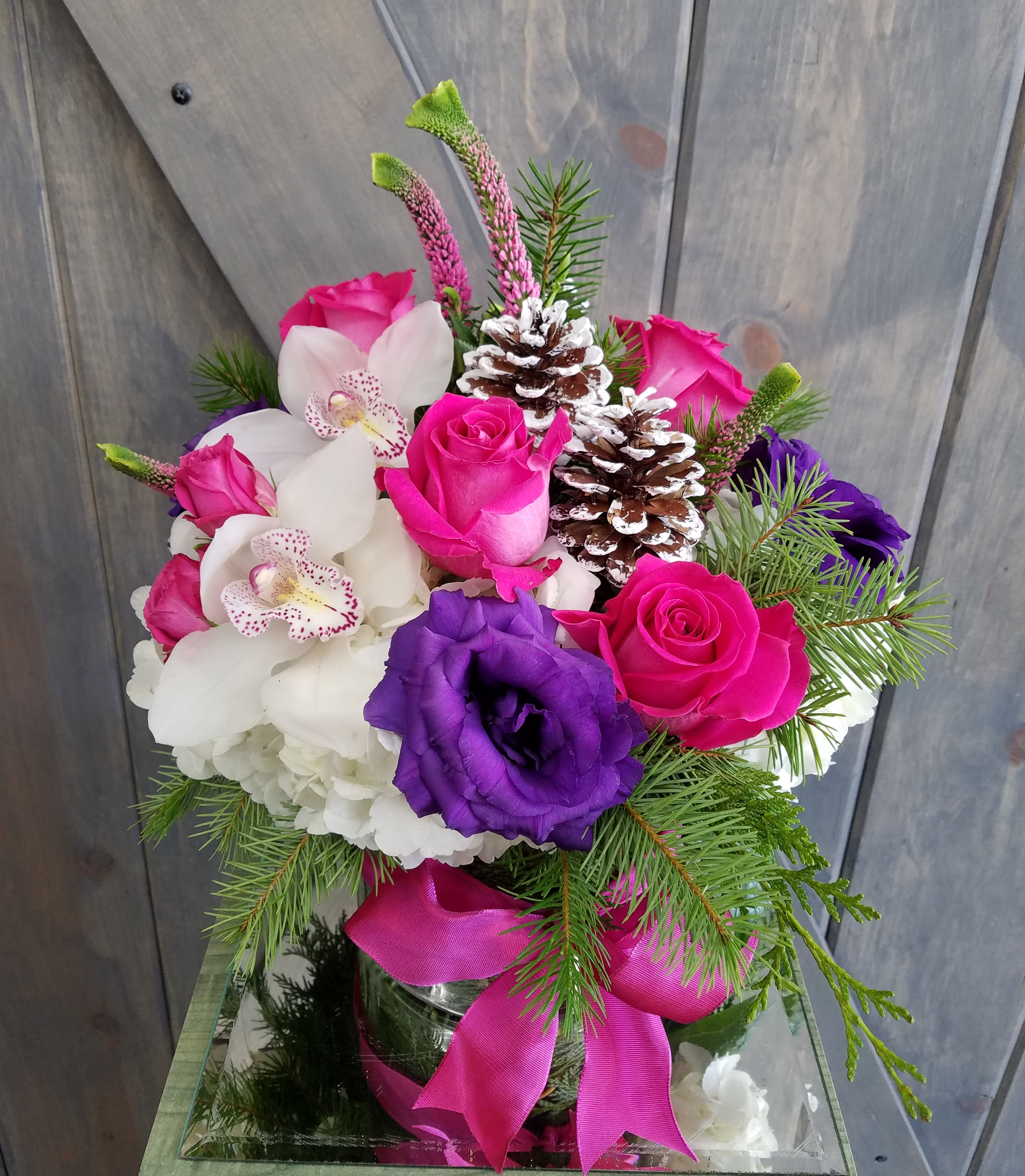 Vibrant Winter - This design is for your non traditional winter celebration. Our design includes Christmas greens, Hot Pink roses, Hot Pink Spray Roses, Purple Lisianthus, White cymbidium orchids, Pink Veronica, whtie hydrangea, and Snowy pine cones