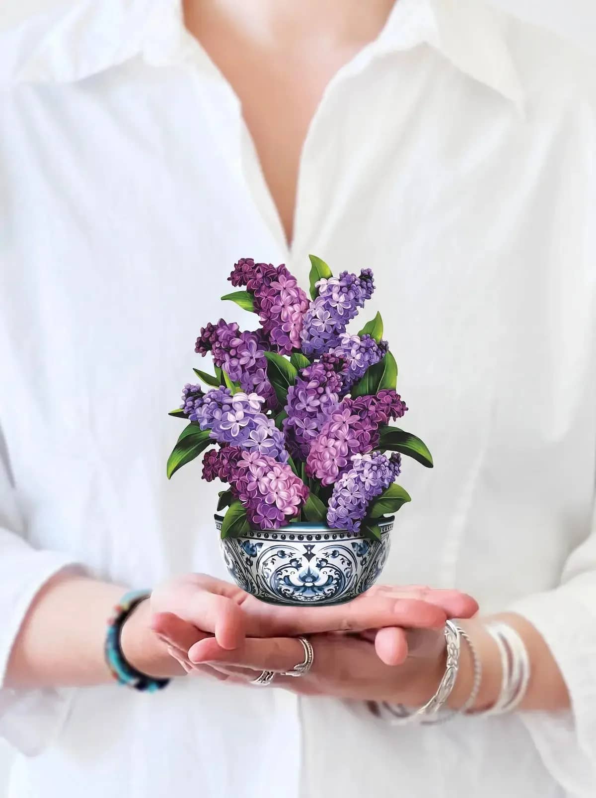 Mini Garden Lilacs Pop-Up Card - Discover the beauty of spring with our Mini Garden Lilacs pop-up bouquet. Standing just below 6 inches, this charming display features lush greenery and luminous purple hues, perfect for any setting. Enjoy the elegance of lilacs without the fleeting blooming season or harsh fragrance. Symbolizing love, acceptance, and confidence, this mini bouquet is an ideal gift for any occasion. Elevate your decor with FreshCut Paper's exquisite designs. Available now!  Measures 6” high by 5” across