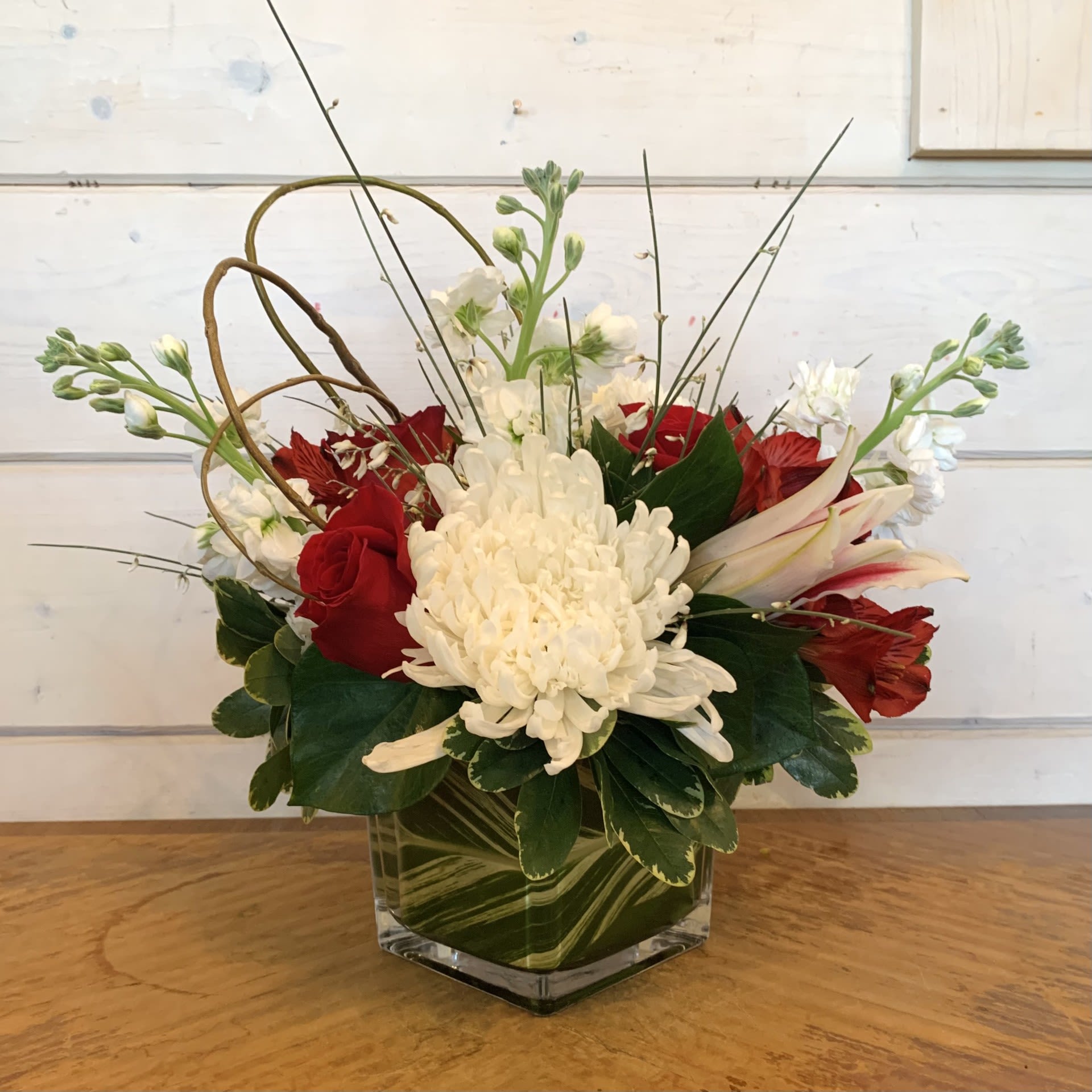 As You Wish - Red roses and stargazer lilies with fragrant white stock, and white crysanthemums in a leaf-lined 5" glass cube with a curly willow loop, red alstroemeria, and seasonal filler flowers. Approximately 10" wide and 10" tall. 