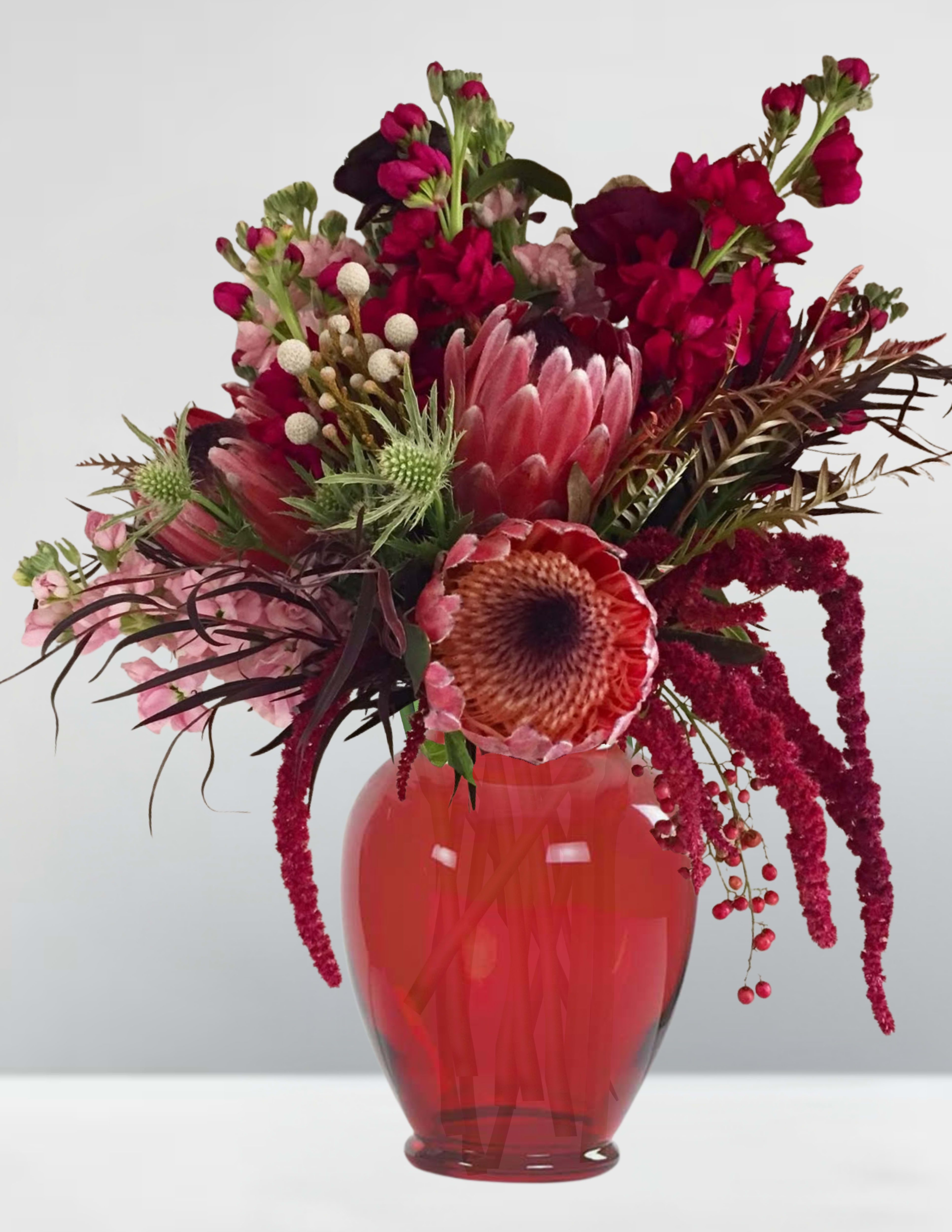 Enchanted stems  - Enchanted Stems is a spellbinding fusion of nature’s wild elegance and mystical charm. Each bloom whispers a secret, each stem curves with intention, as if plucked from a forest where magic lingers in the morning mist. Deep crimson proteas rise like crowned guardians, surrounded by cascading amaranthus that drips like enchanted wine. Thistle-like greens and berry-laced sprigs add texture and intrigue, while snapdragons and fern-like foliage weave a tale of forgotten gardens and twilight rituals.  This arrangement doesn’t just sit in a vase—it casts a mood. It’s the kind of bouquet you’d expect to find in a moonlit greenhouse, tended by someone who knows the language of petals and shadows.