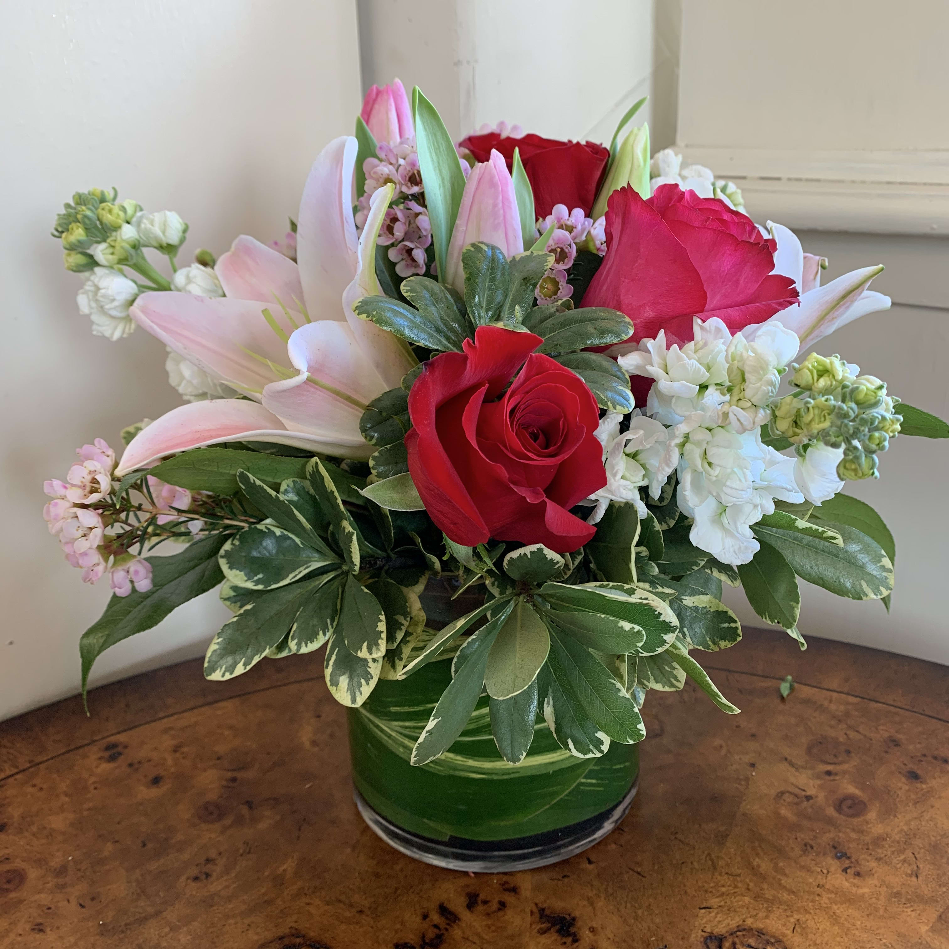 Love Me - Mixed red and hot pink roses, pink lilies, pink tulips, and white stock arranged with pink wax flower for filler in a leaf lined small gather vase. Approximately 10" tall by 10" wide. 