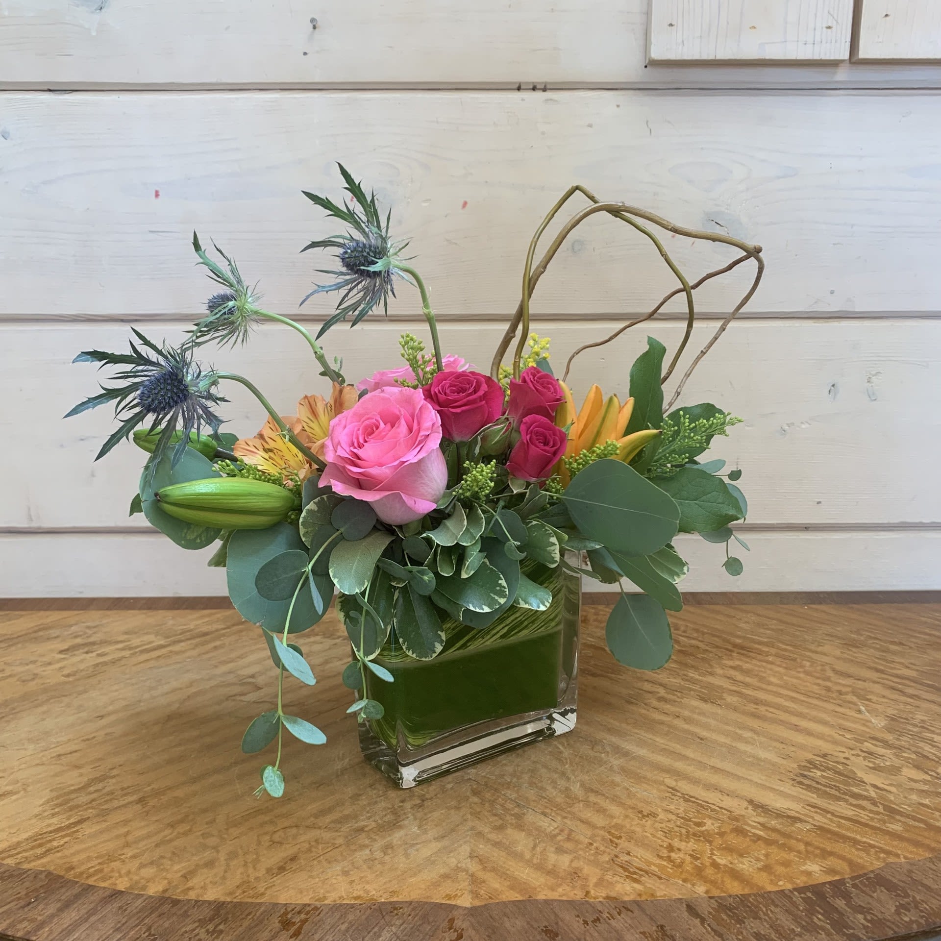 Midsummer Mosaic - Pale pink roses, hot pink spray roses and orange lilies arranged in a narrow mini flat rectangle with curly willow looped, dramatic blue thistle, orange alstroemeria and yellow solidago with eucalyptus greens. Approximately 10" tall, 10" wide and 6" deep.