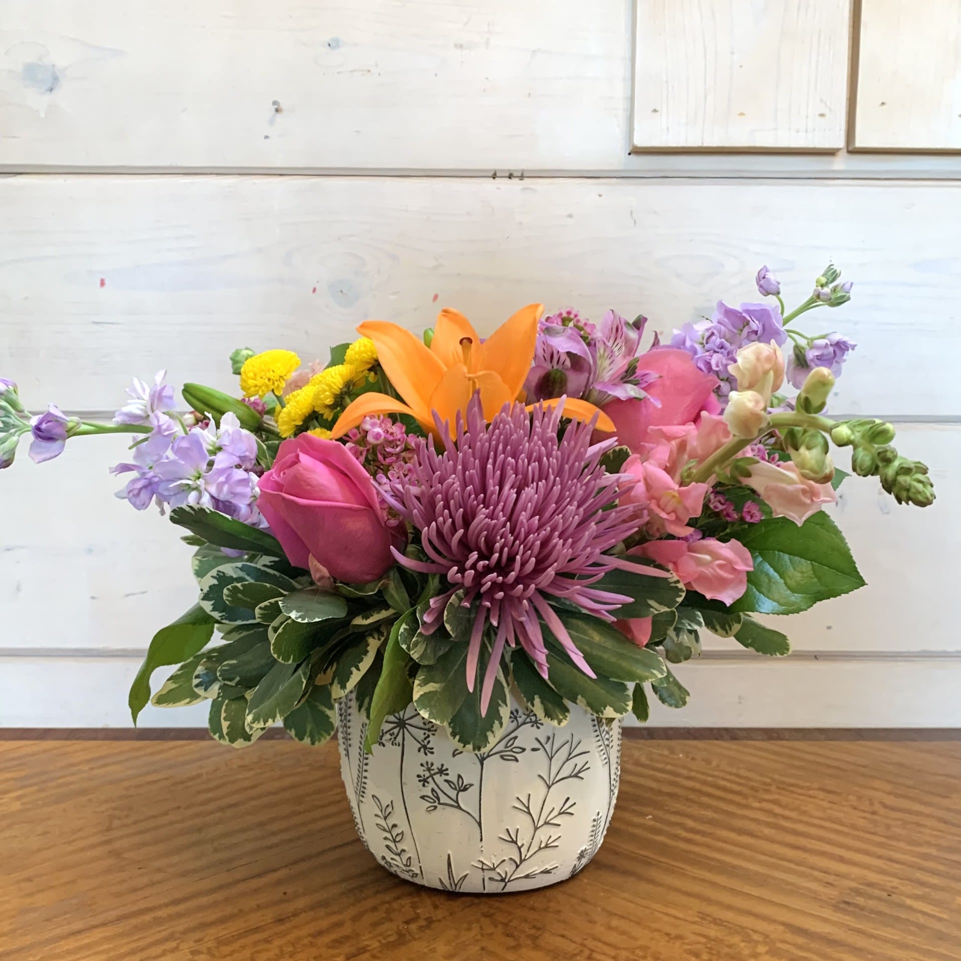 A Cheerful Spring Bouquet - An orange lily, pink roses, pink snapdragons, lavender stock, yellow and green chrysanthemums, and purple alstroemeria arranged in a small white pot embossed with black wildflowers, with seasonal filler flower. Approximately 12" tall and 12" wide.