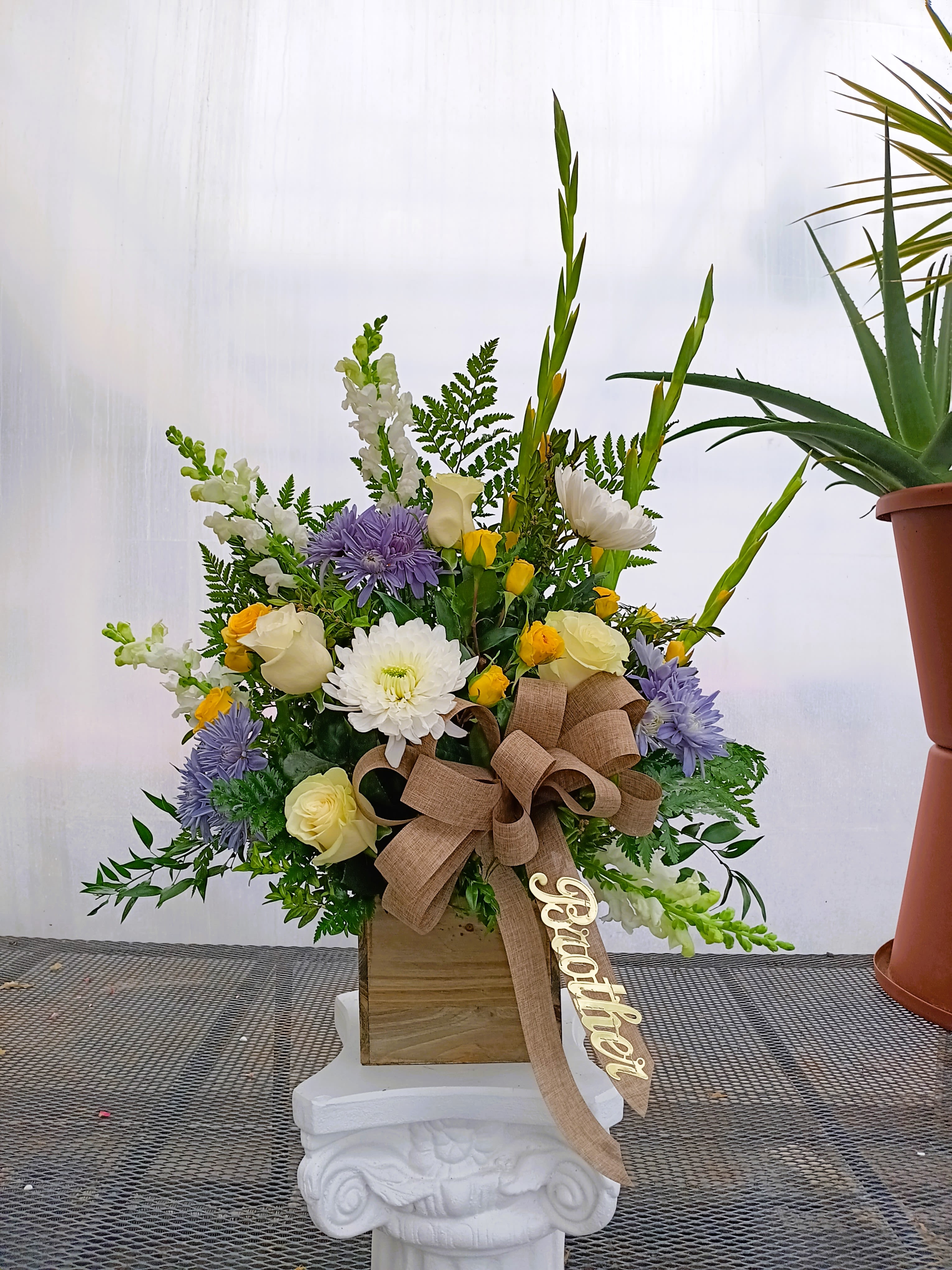 Funeral Traditional - Rustic Remembrance Tribute - Celebrate life with this elegant and traditional funeral arrangement, crafted with bright yellows, serene whites, and calming blues. Symbolizing warmth, peace, and reflection, this piece features a harmonious blend of seasonal blooms thoughtfully designed to honor a life well lived. Perfect for conveying comfort and support, this piece reflects a life of love, light, and cherished memories.  The arrangement is accented with a rustic burlap country ribbon, adding a heartfelt, personal touch. The ribbon bears the loving script: "Brother," a timeless tribute to his enduring legacy. Script and colors can be customized to suit any color scheme.