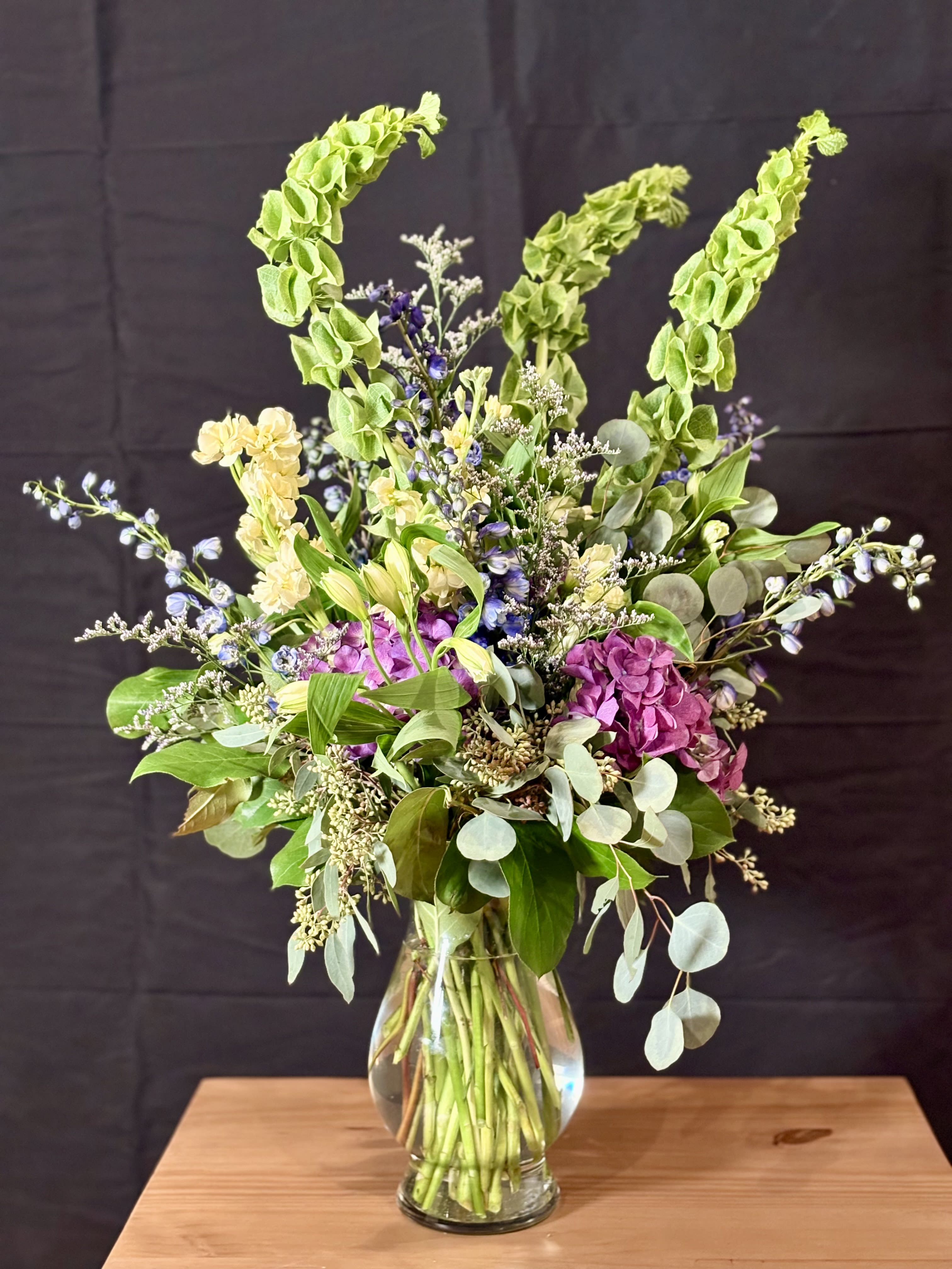 Modern Medow - This fresh, garden-style arrangement is full of movement and natural elegance. Featuring sculptural green bells of Ireland rising above lush hydrangea in rich berry tones, it’s accented with soft yellow stock, airy blue delphinium, and delicate filler flowers for texture. Eucalyptus and layered greenery add depth and a relaxed, organic feel, all gathered in a clear glass vase that highlights the natural beauty of the stems. Vibrant yet refined, this design feels effortlessly gathered, perfect for brightening a space or making a graceful, modern statement.