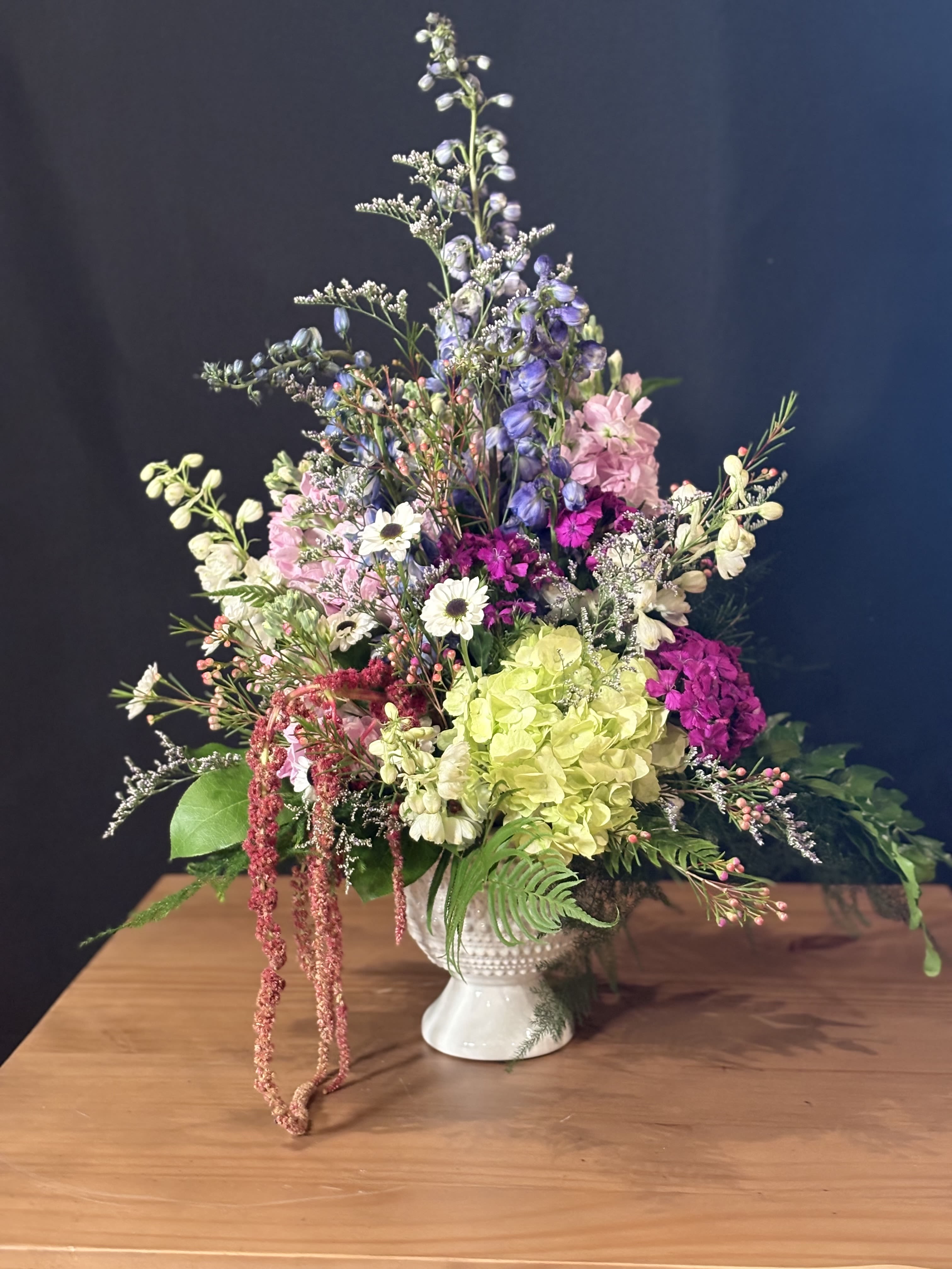 Wildflower Dreams - This lush, garden-inspired arrangement is a celebration of texture, color, and natural movement. Designed in a classic pedestal vase, it features an abundant mix of seasonal blooms, soft hydrangea, airy delphinium, delicate daisy-like flowers, and romantic stock, layered with wisps of waxflower and fresh greenery. Cascading amaranthus adds an elegant trailing touch, while varied heights and tones create a graceful, meadow-like silhouette. Full, expressive, and artfully arranged, this piece makes a stunning statement for celebrations, heartfelt gestures, or simply bringing beauty into any space.