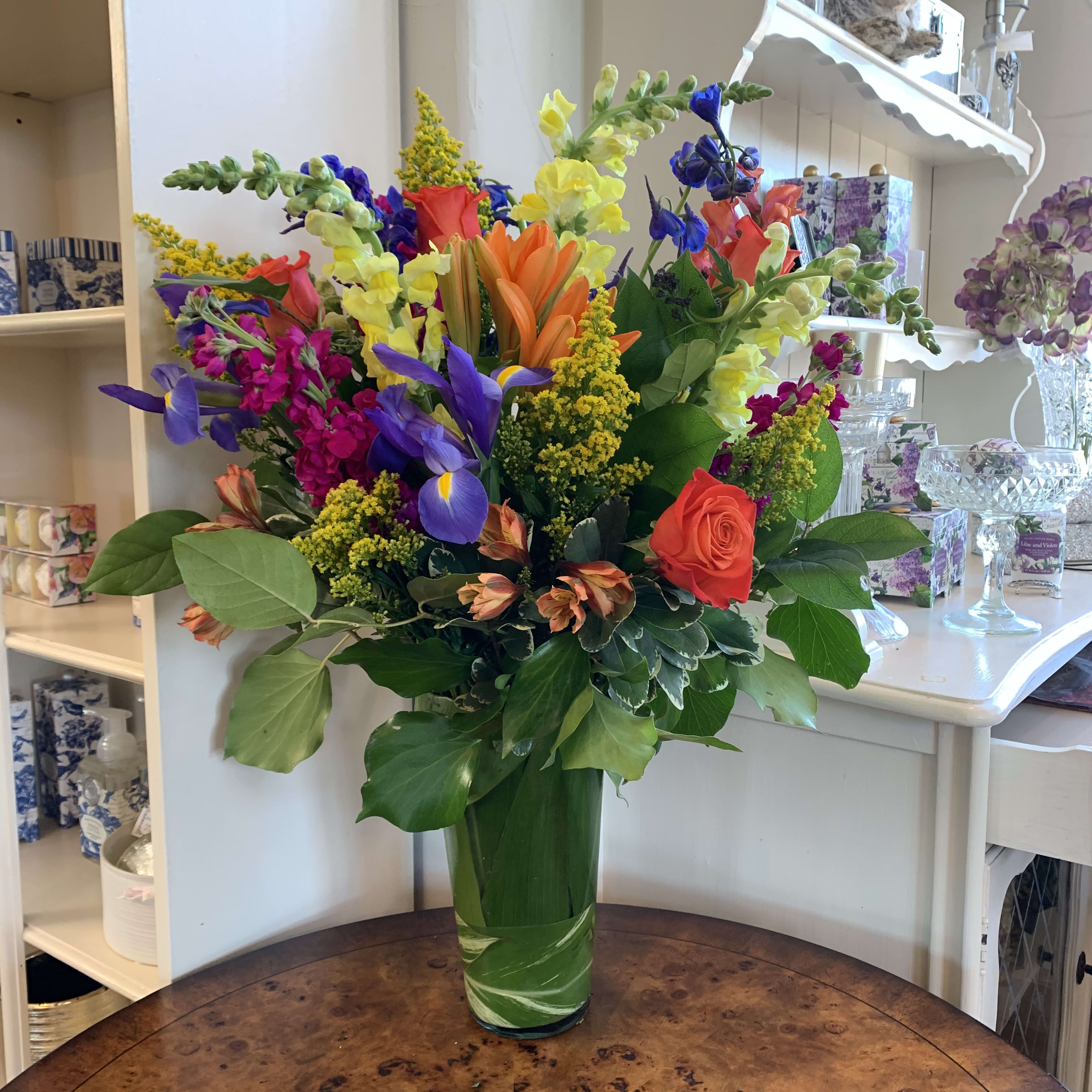 Bright Horizons  - orange roses, orange lilies, yellow snapdragosn, magenta stock, iris, blue delphiniam, and orange alstromaria arranged with solidago in a leaflined french vase. Approximatly 24in tall and 20in wide. 